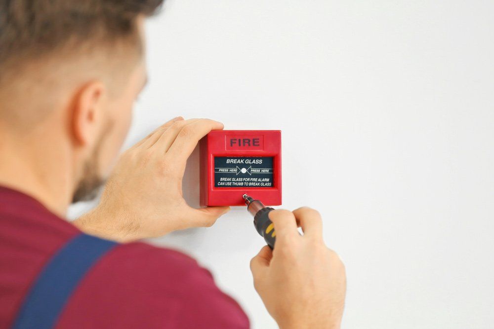 Man Installing a Red Fire Alarm on a White Wall With a Screwdriver — Hunter Fire & Safety in Edgeworth, NSW