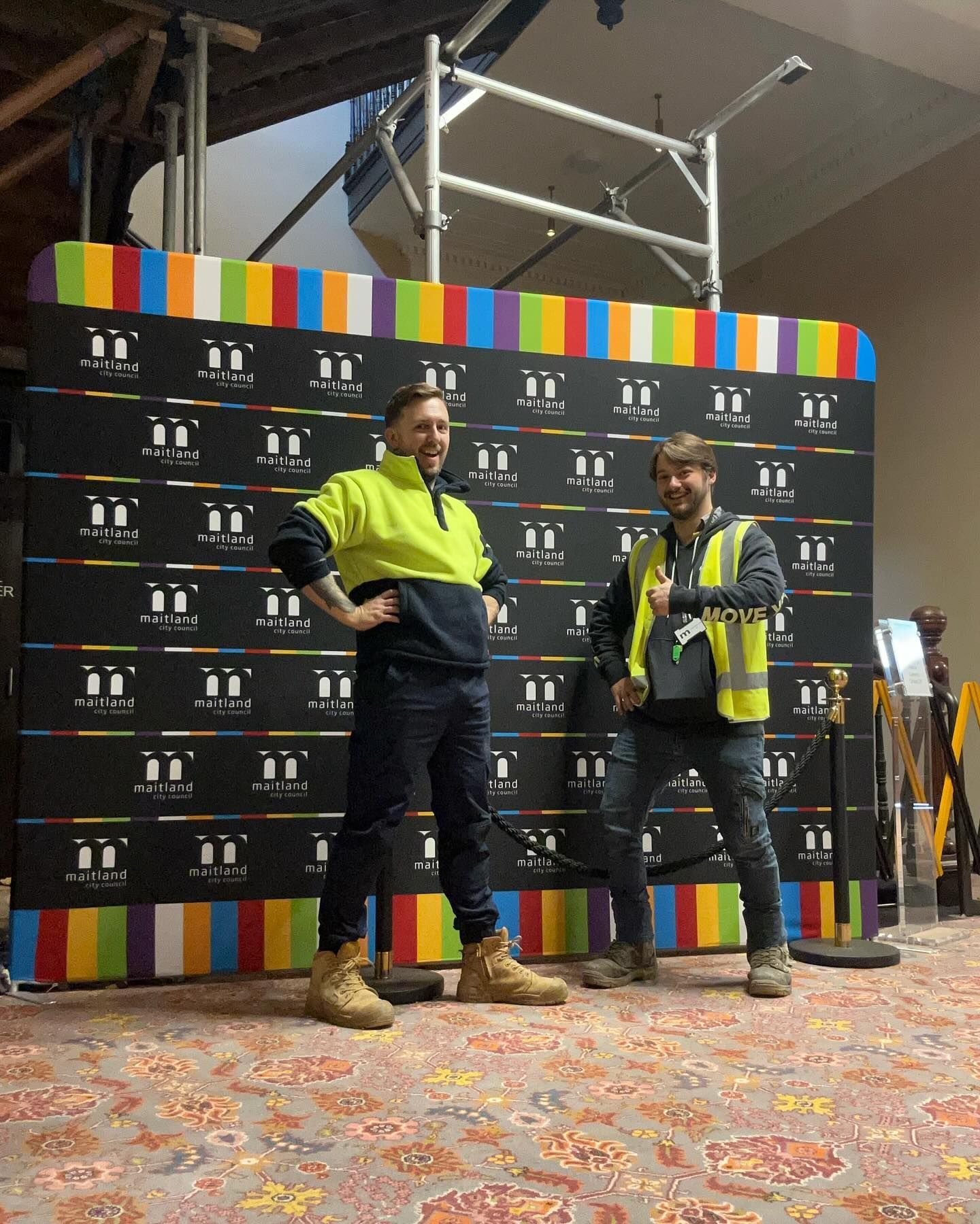 Two Men Pose in Front of a Colorful Backdrop. One Wears a Green Safety Vest — Hunter Fire & Safety in Lake Macquarie, NSW
