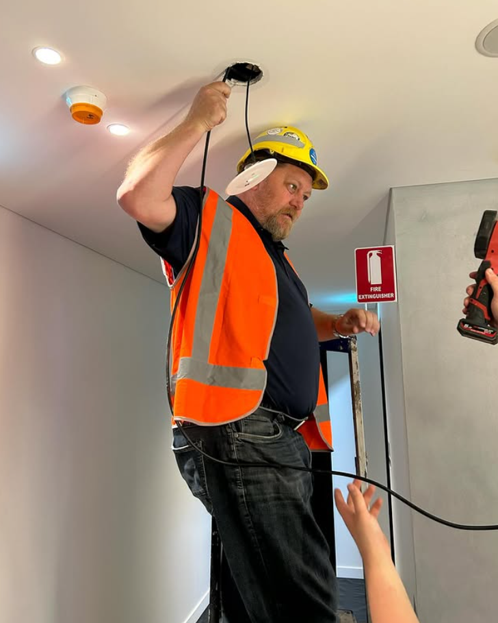 Man in safety vest, hard hat working on ceiling wiring. Hallway setting, fire extinguisher visible.
