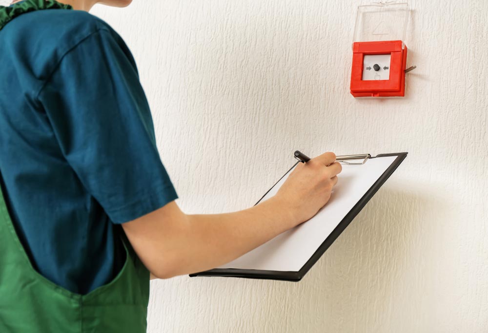 Worker With Clipboard Near Modern Fire Call Point On Wall — Hunter Fire & Safety in Central Coast, NSW