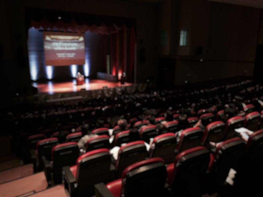 A blurred image of people sitting in an auditorium watching a presentation