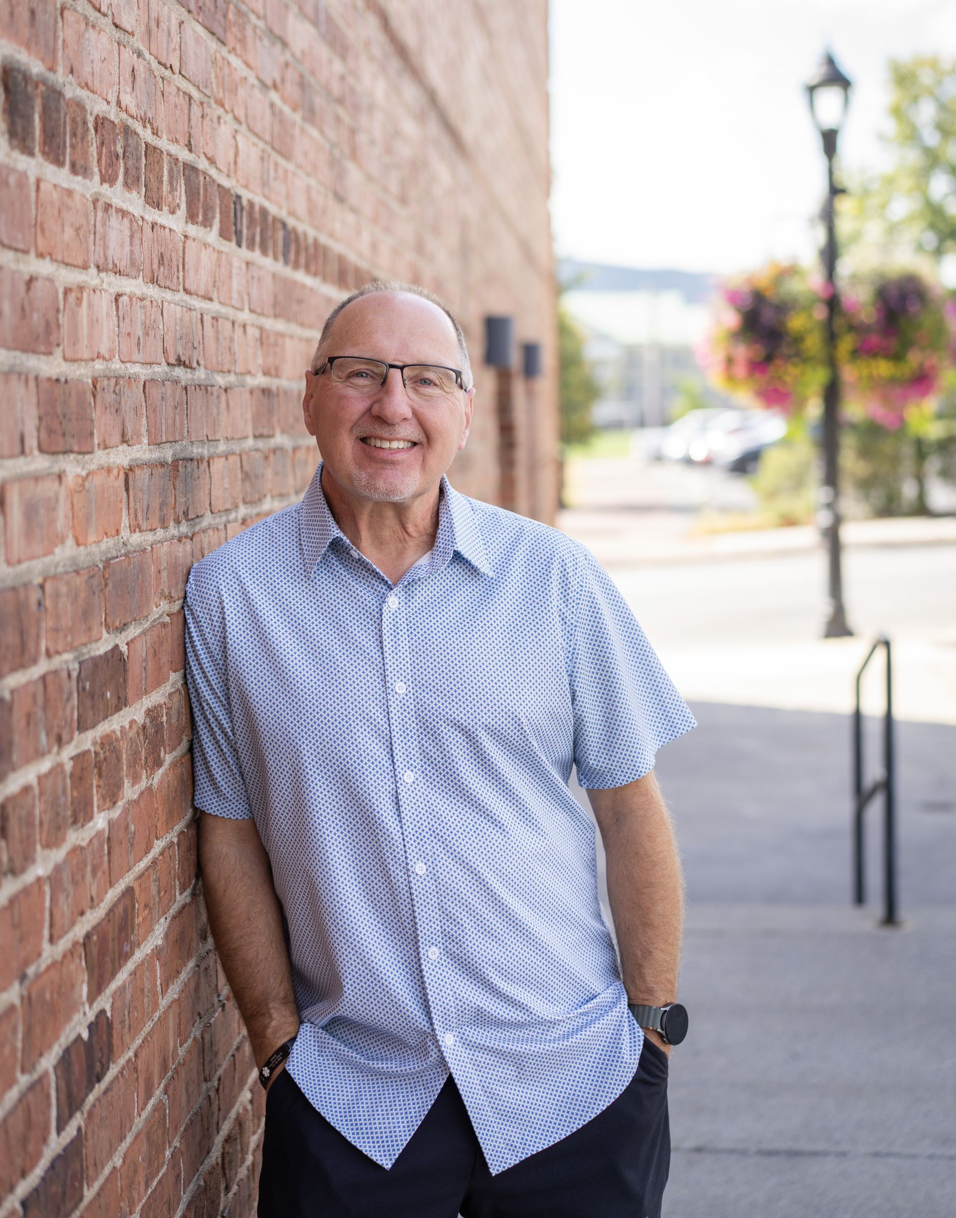 Man leans against brick wall, smiling. He wears a patterned button-up shirt and black pants, hands in pockets, outside.