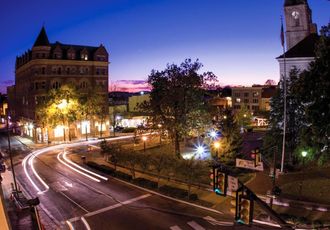 Nighttime cityscape with illuminated buildings, streetlights, and blurred car trails under a twilight sky.