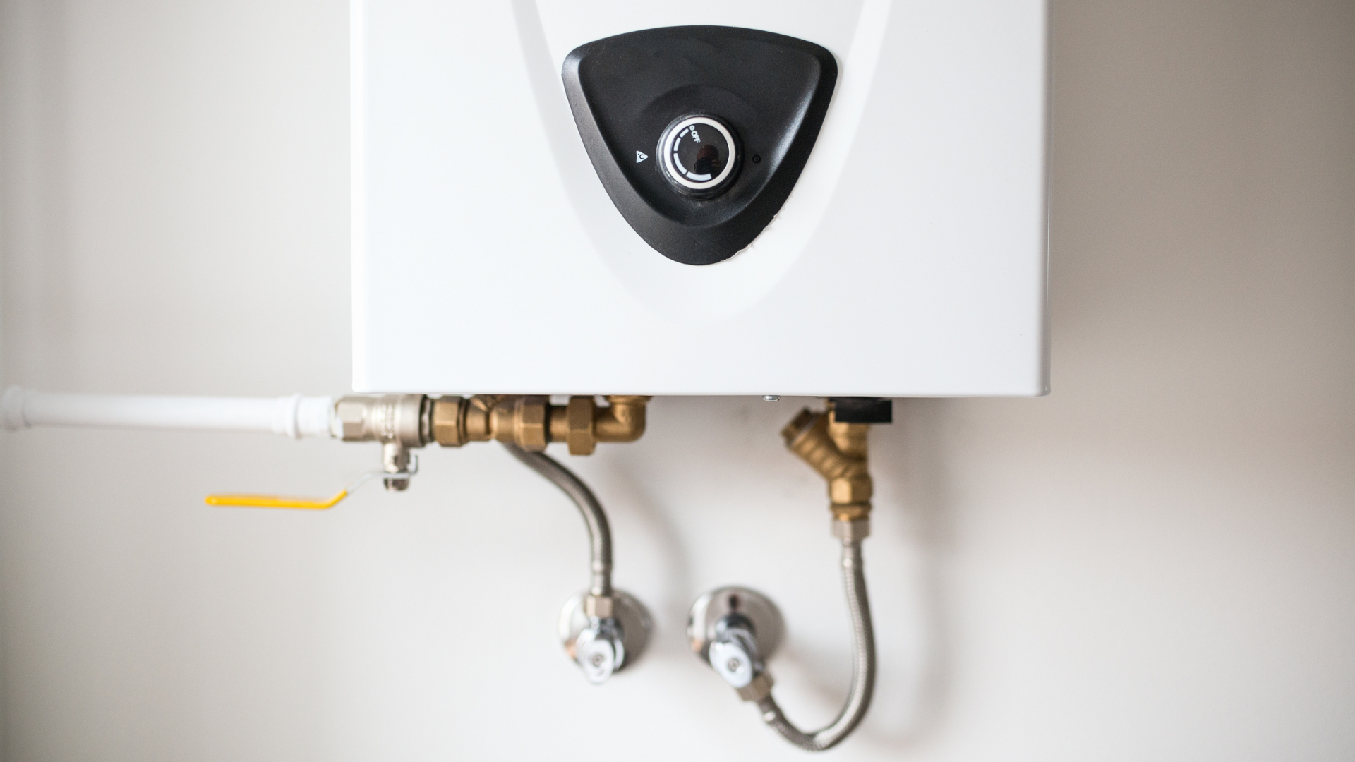 A white tankless water heater mounted on a wall with metal plumbing pipes and yellow valves below.