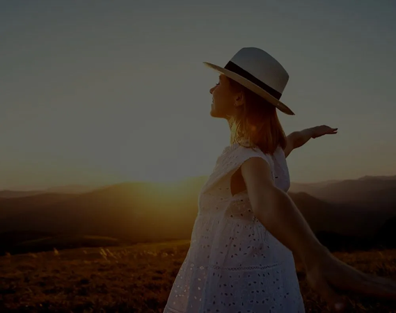 A woman in a white dress and hat is holding a man 's hand in a field at sunset.