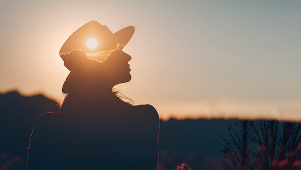 A woman in a hat is standing in a field at sunset.