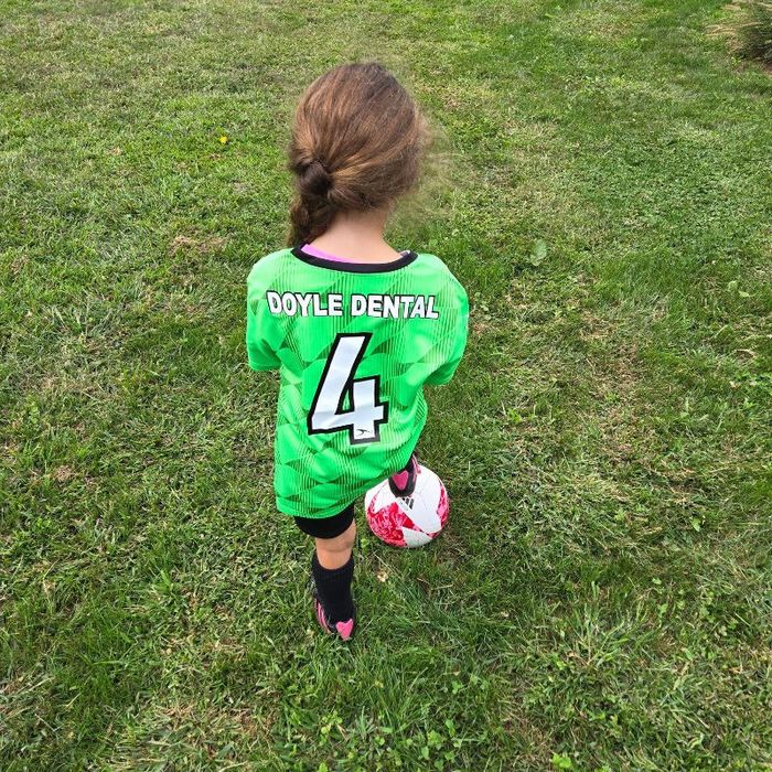 A little girl wearing a green jersey with the number 4 is holding a soccer ball.