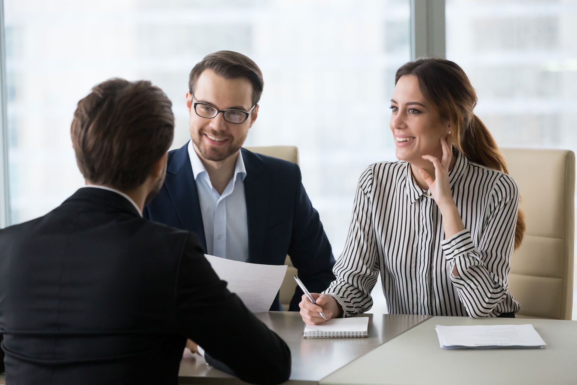 Three people in suits at a table, two smiling interviewing a man with documents in an office.