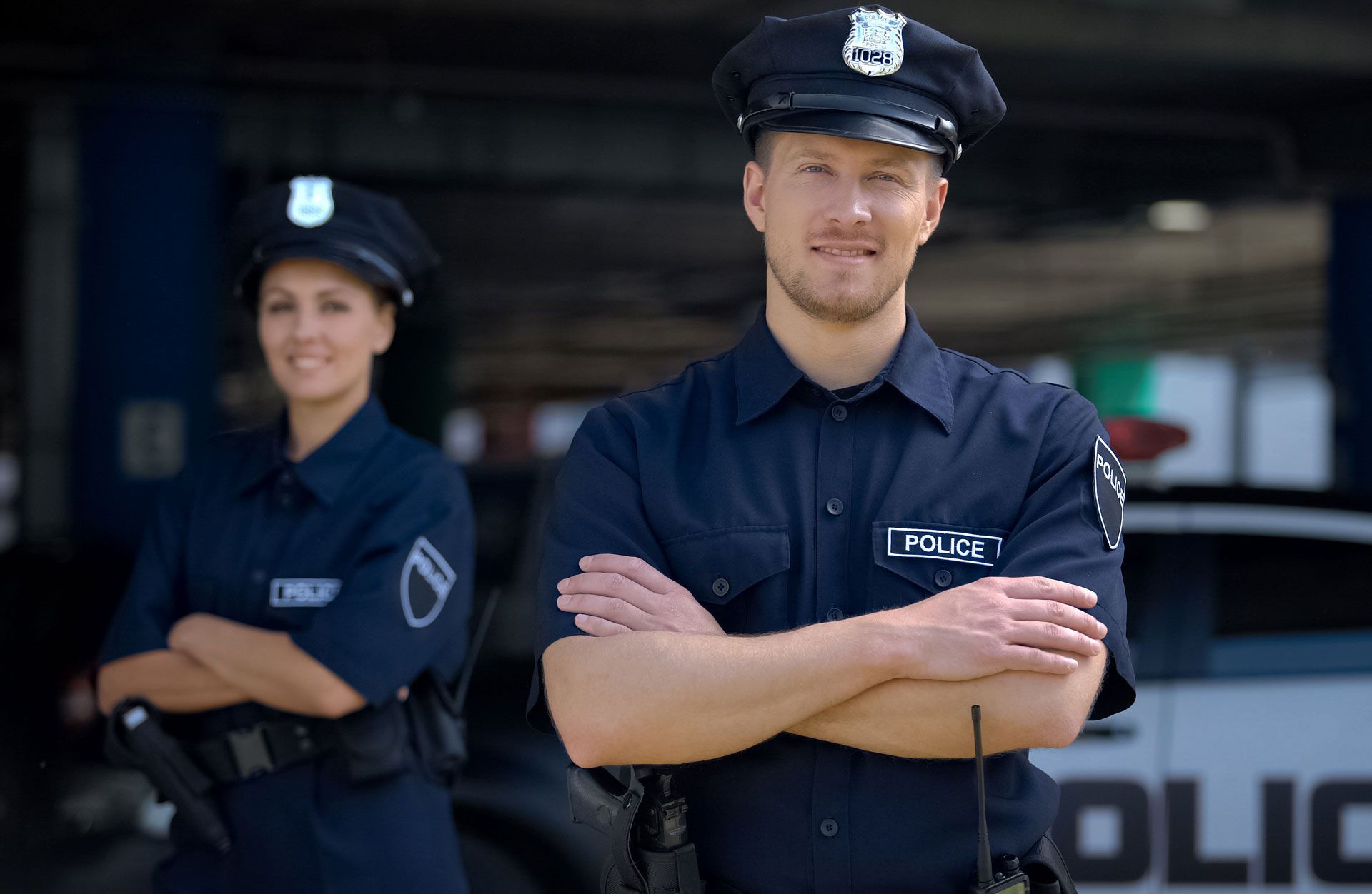 Two police officers in uniform standing with arms crossed, smiling near a police car.