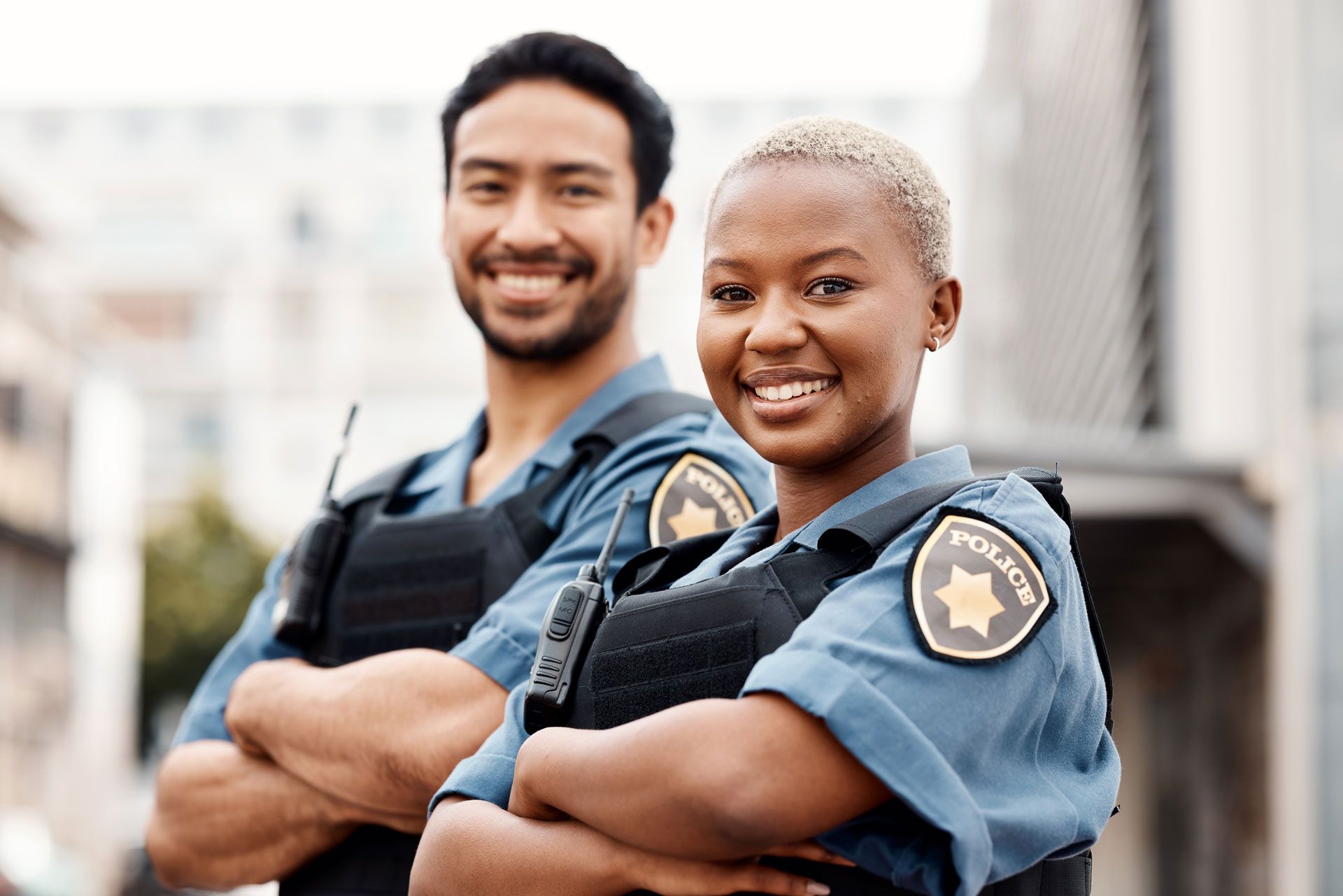 Two smiling police officers, an Asian man and Black woman, arms crossed. They wear uniforms outdoors.