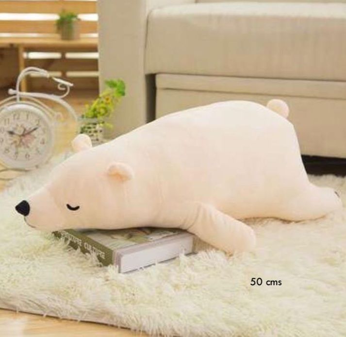 Almohada de peluche en forma de oso polar, de color crema, tumbado sobre una alfombra cerca de un sofá.