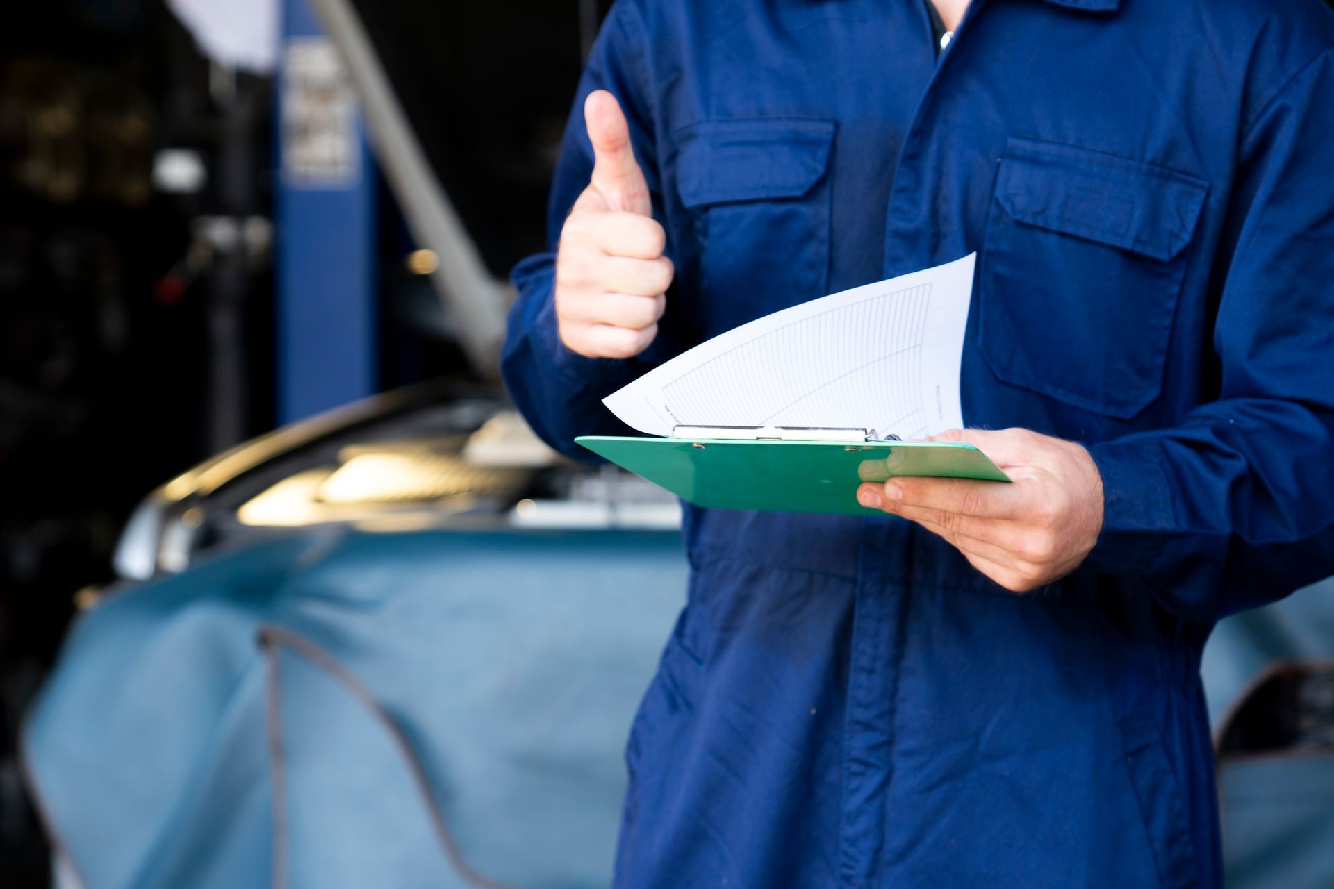 Mechanic standing, thumbs up and holding clipborad with blur garage in the background.