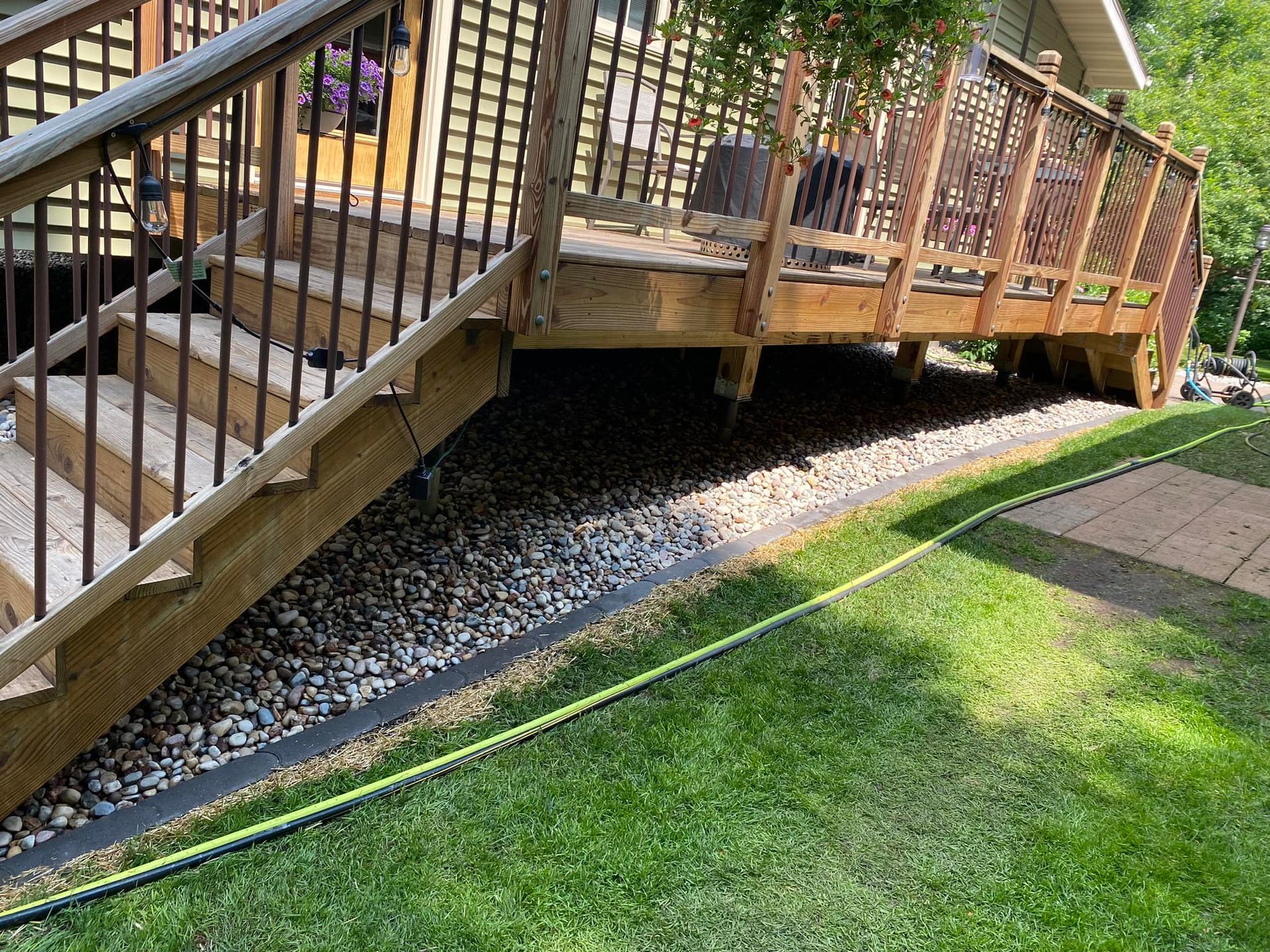 A wooden deck with stairs leading up to it and a hose in the grass.