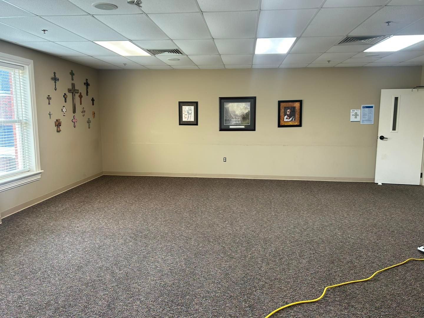Empty room with brown carpet, beige walls, and framed pictures. A crucifix is on the wall.