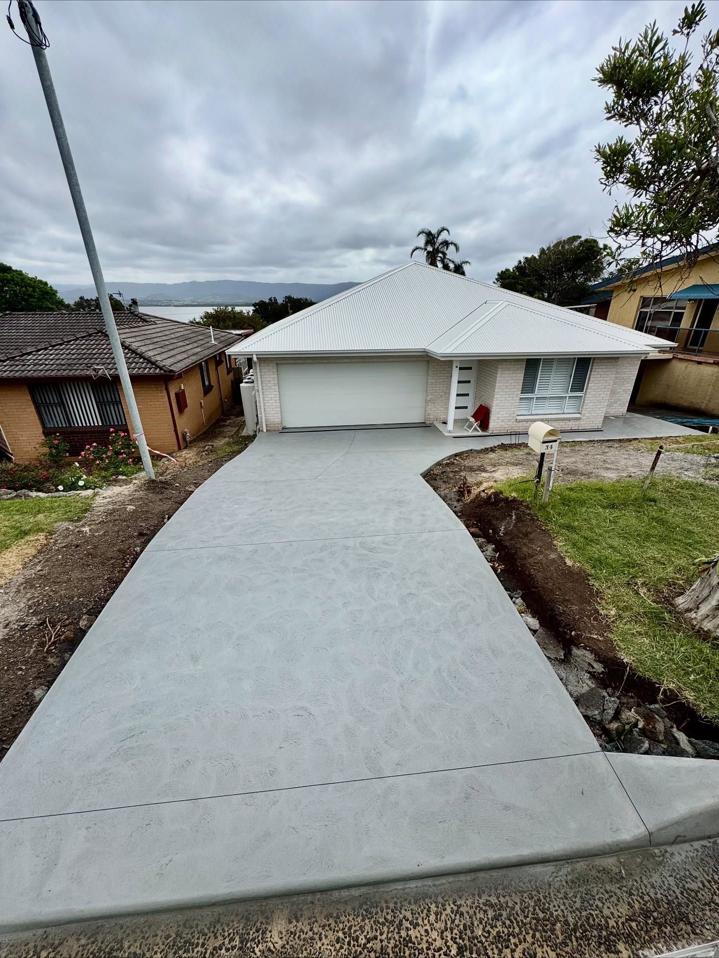 A wide, newly poured concrete driveway leads up to a light-colored, single-story house under a cloudy sky. — JDB Concreting in Horsley, NSW