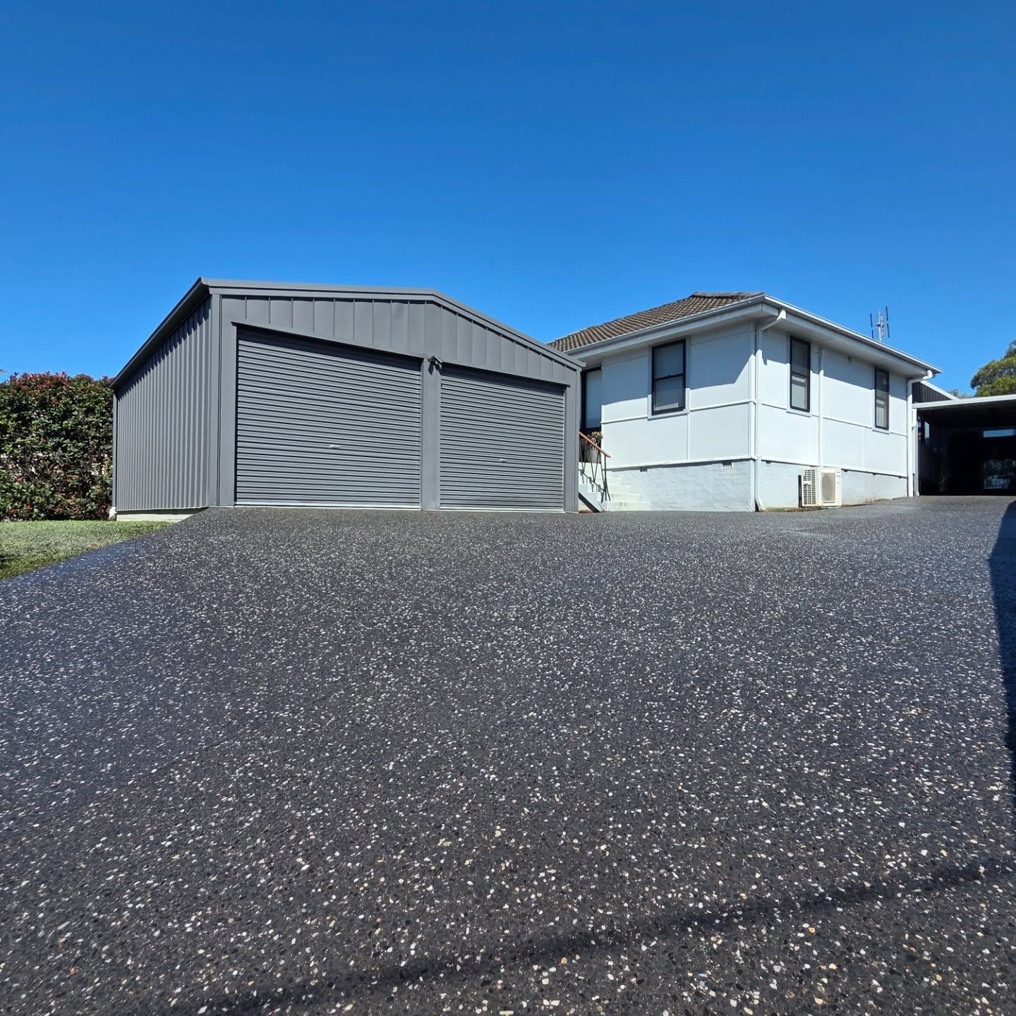 A white house with a gray metal double garage and a large, speckled dark asphalt driveway under a clear blue sky. — JDB Concreting in Horsley, NSW