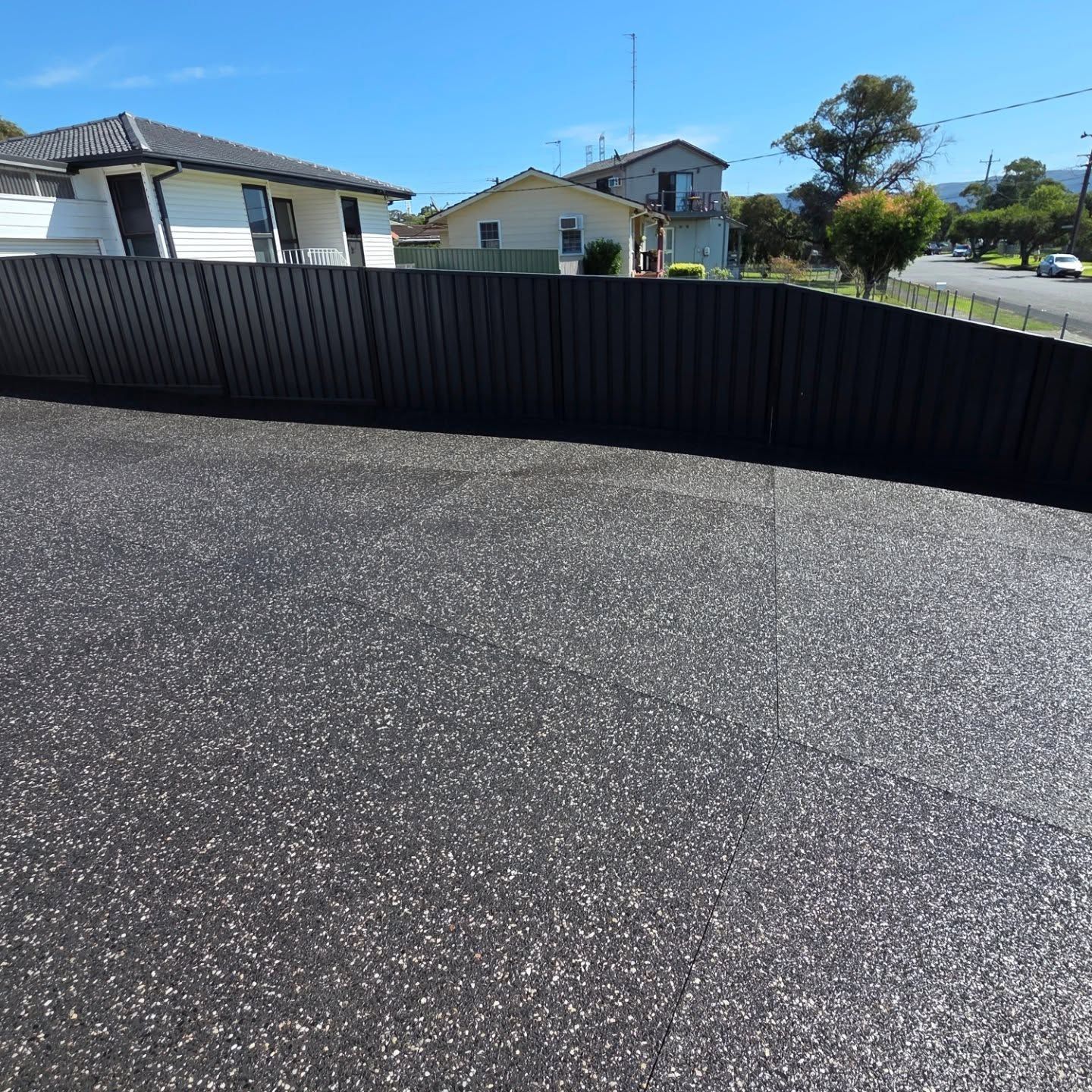 A modern black metal fence bordering a gray aggregate driveway in front of suburban houses under a clear blue sky. — JDB Concreting in Woonona, NSW
