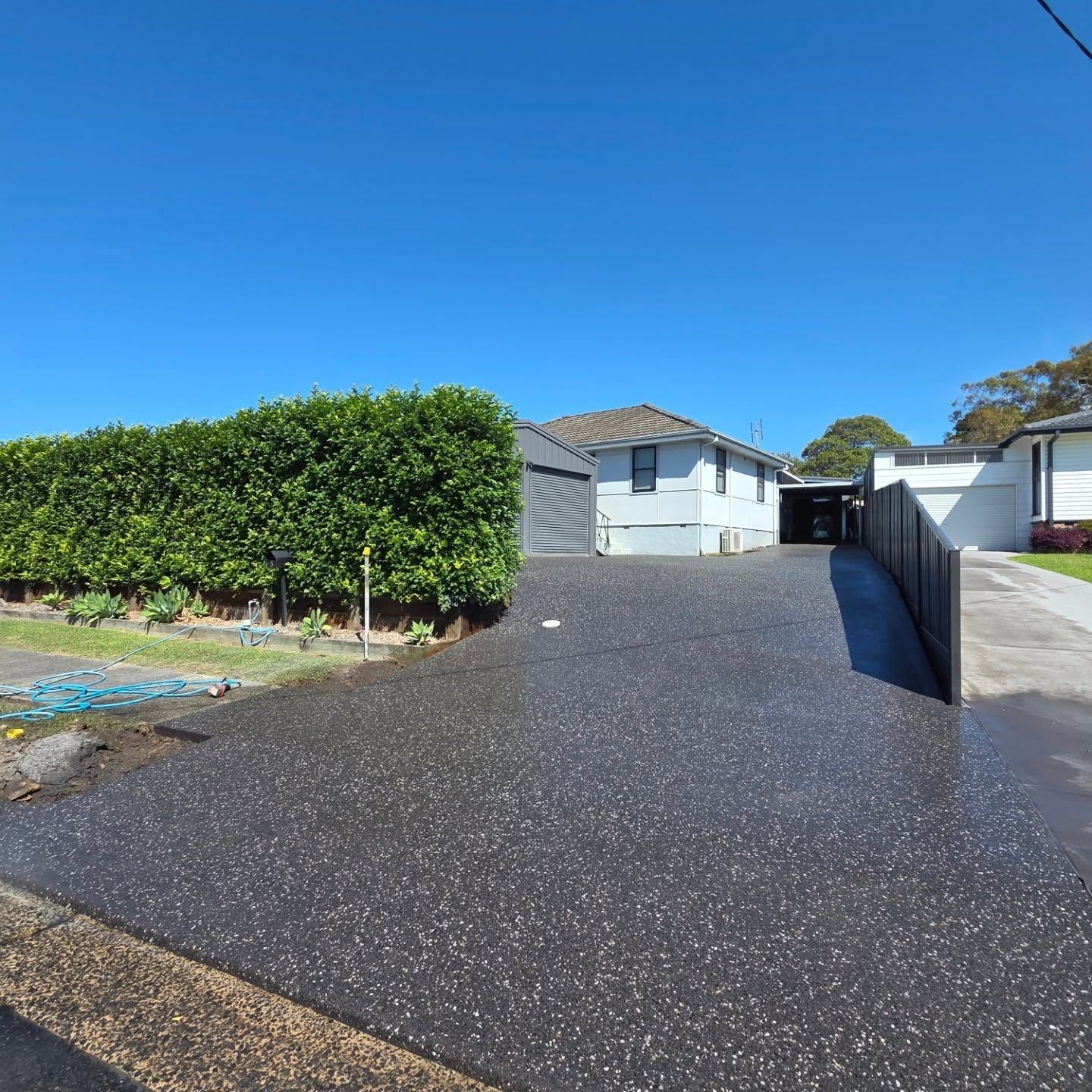 A driveway with a dark, speckled finish leads toward a light-colored house beside a tall green hedge under a clear sky. — JDB Concreting in Horsley, NSW
