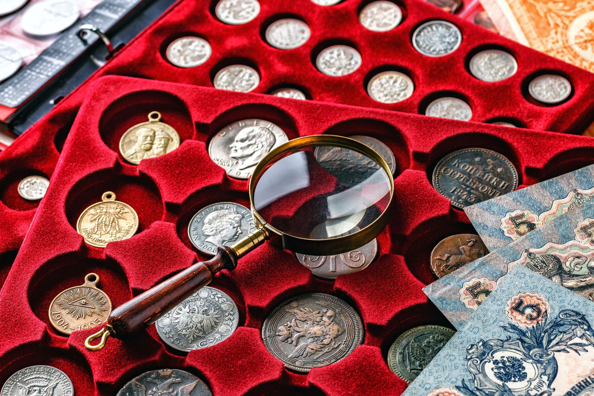 Magnifying glass on red case holding assorted antique coins and paper currency.