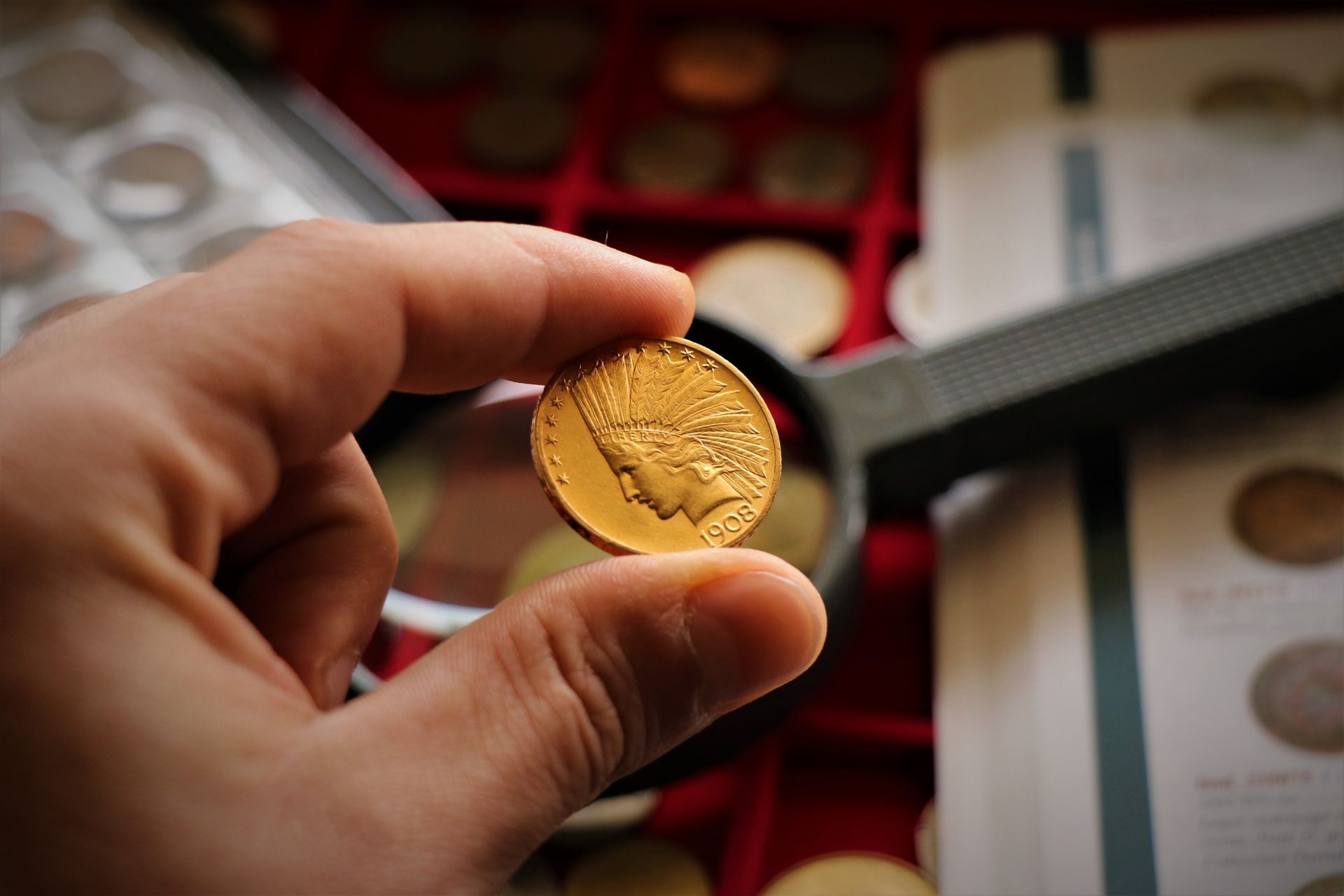 Hand holding a gold Indian Head coin above a coin collection tray and magnifying glass.
