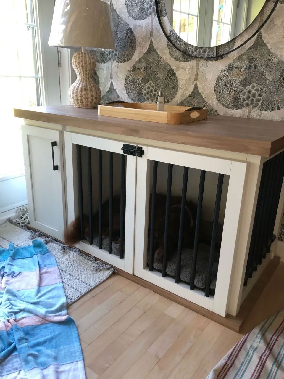 Dans l'entrée d'une maison, une cage pour chien en bois, de style meuble, avec des portes blanches et des barreaux en métal noir, est surmontée d'une lampe.