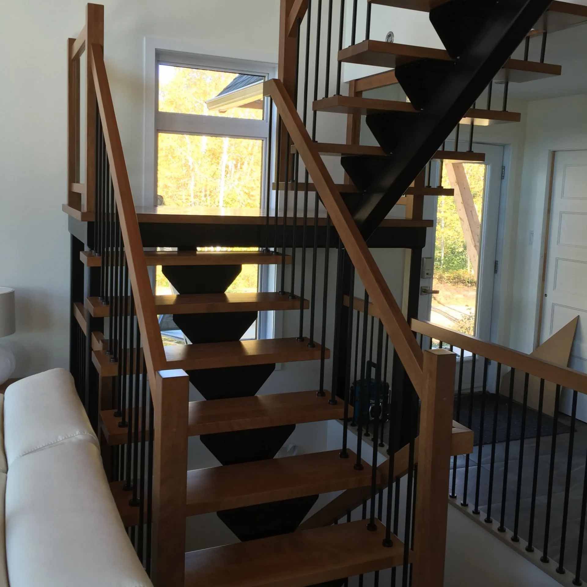Escalier moderne avec marches en bois clair, supports en métal noir et fines rampes verticales dans une pièce baignée de soleil.