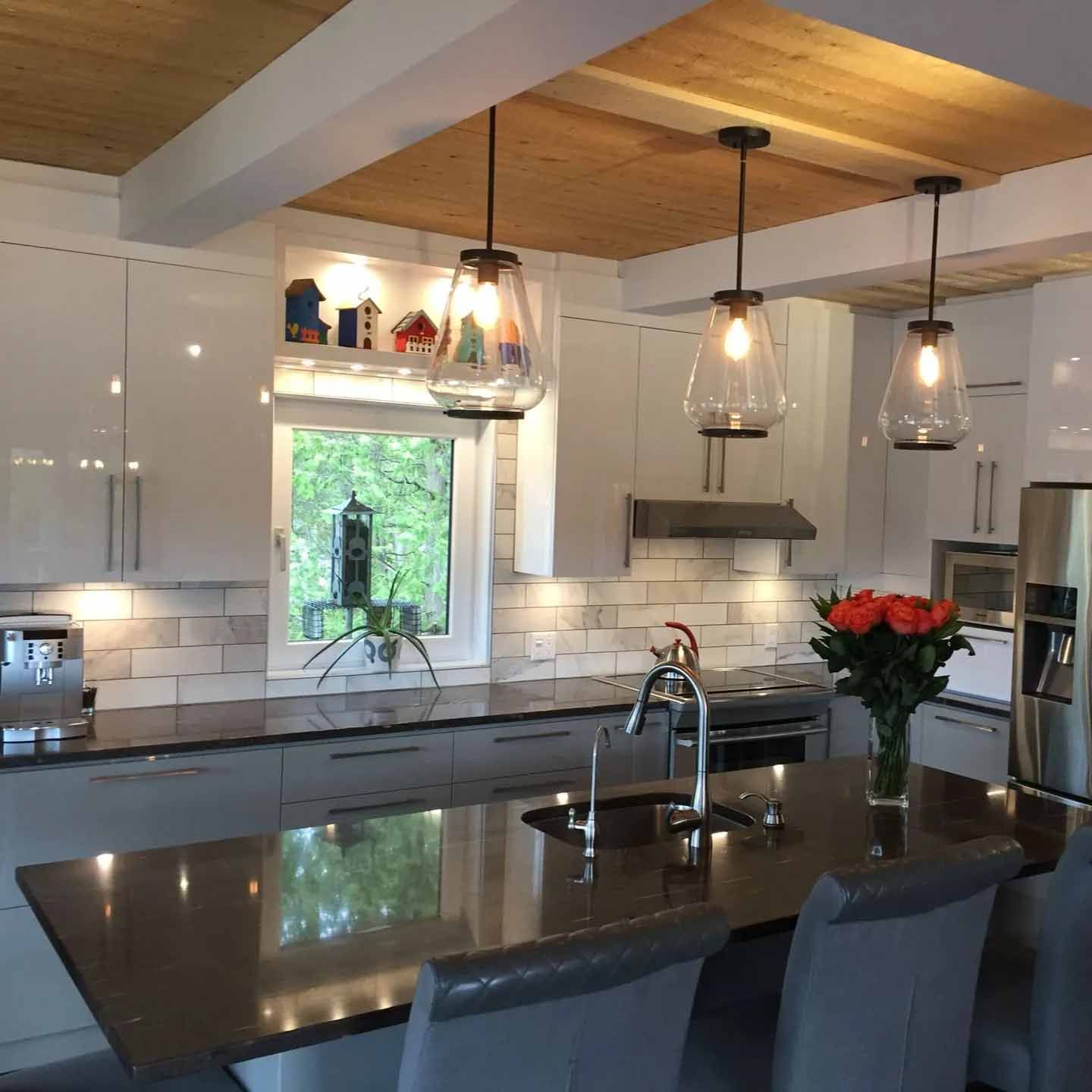 Cuisine moderne avec armoires blanches, plafond en bois, comptoirs foncés, îlot central avec tabourets et luminaires suspendus.