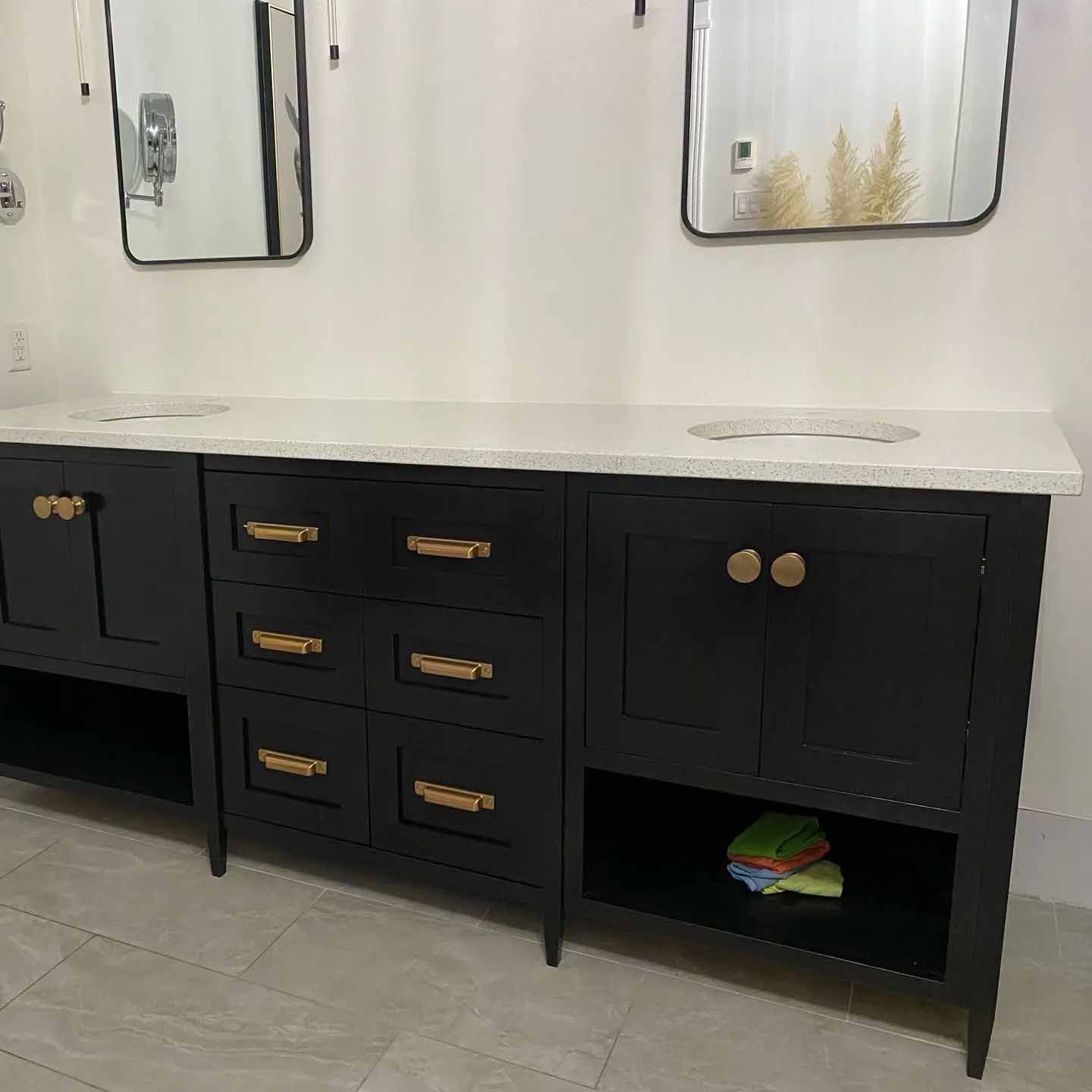 Un meuble-lavabo double pour salle de bain avec des armoires noires, des poignées dorées, des comptoirs blancs mouchetés et deux miroirs rectangulaires.