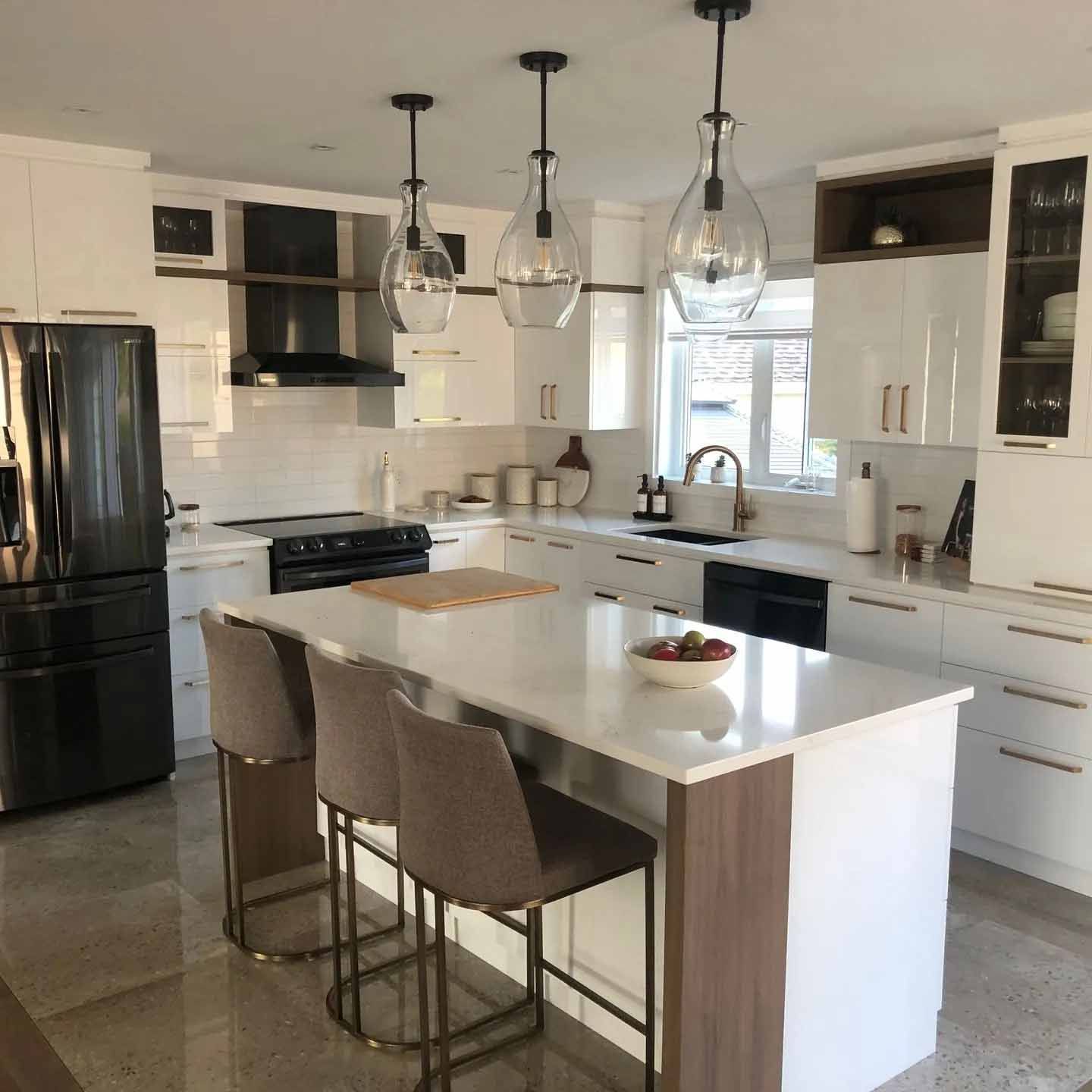 Une cuisine moderne avec des armoires blanches, un grand îlot avec trois tabourets, des appareils électroménagers noirs et trois suspensions en verre.