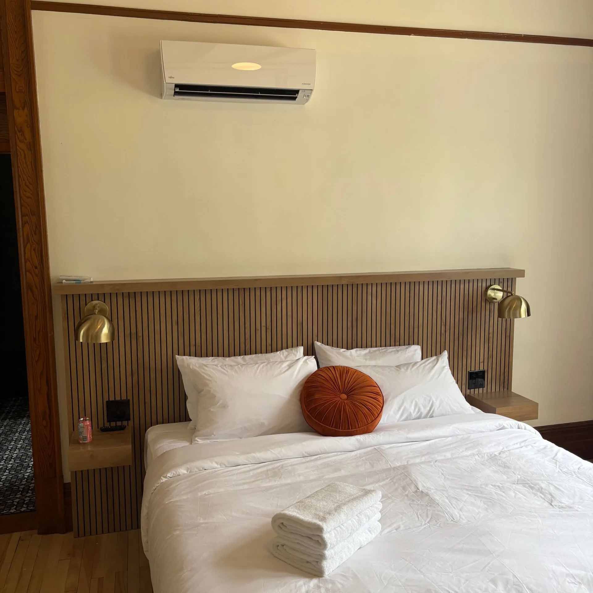 Une chambre avec un lit queen-size doté d'une tête de lit à lattes de bois, de draps blancs, d'un oreiller rond couleur rouille et d'un climatiseur.