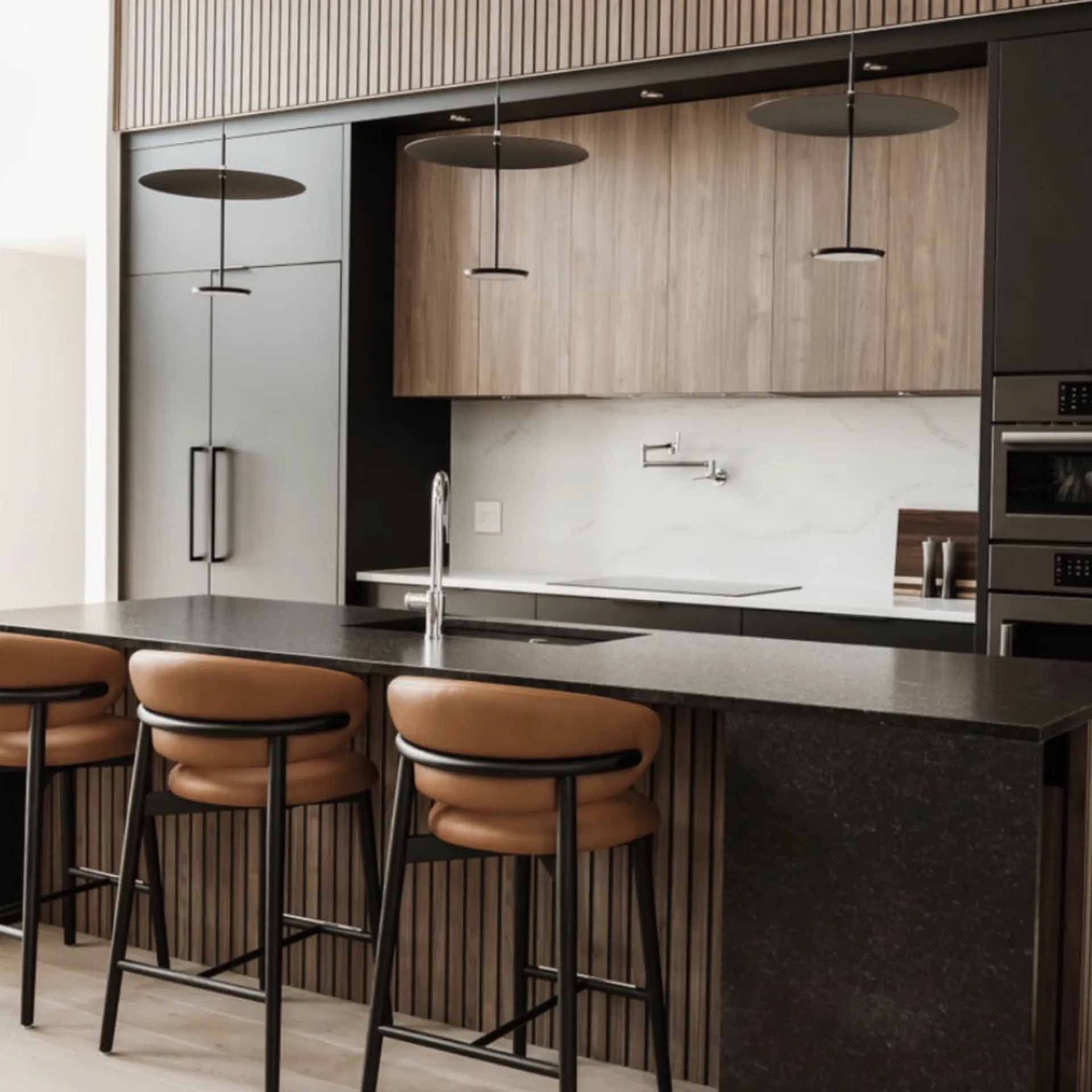 Une cuisine moderne avec un îlot central sombre, des tabourets de bar beiges, des meubles en bois et des suspensions lumineuses noires mates.
