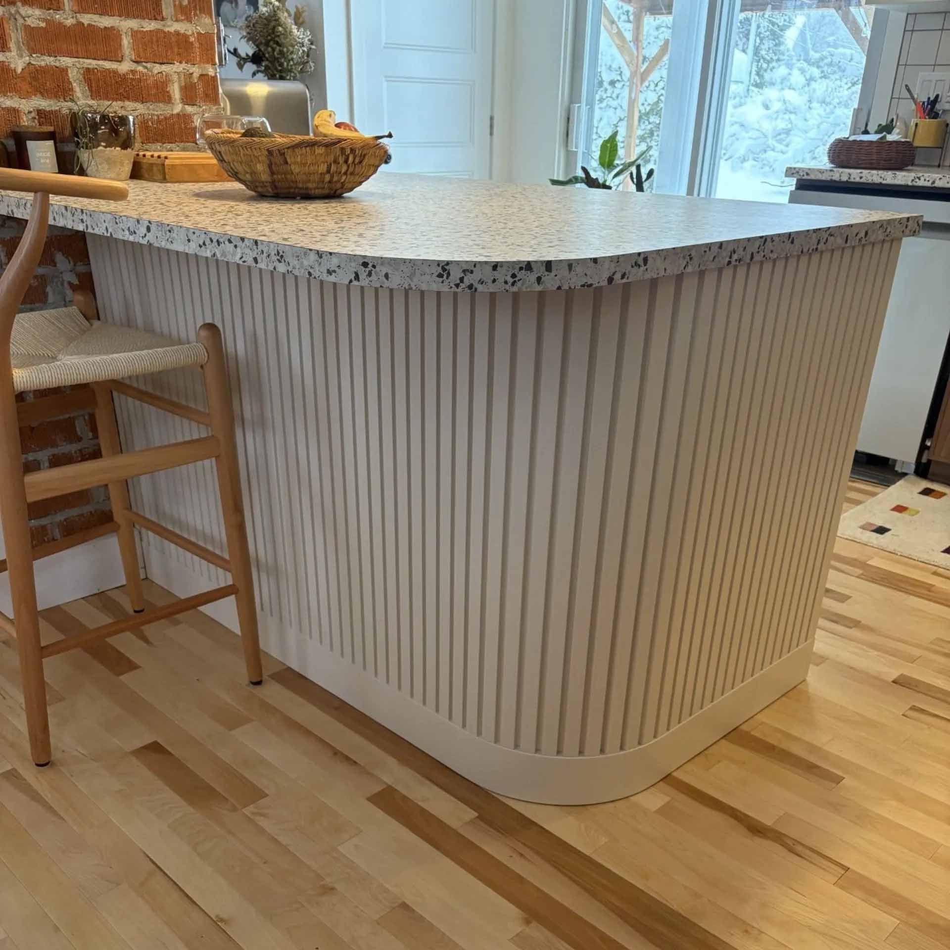 Un îlot de cuisine aux bords arrondis, orné de panneaux cannelés blancs et d'un plan de travail en terrazzo moucheté, est placé à côté d'une chaise.