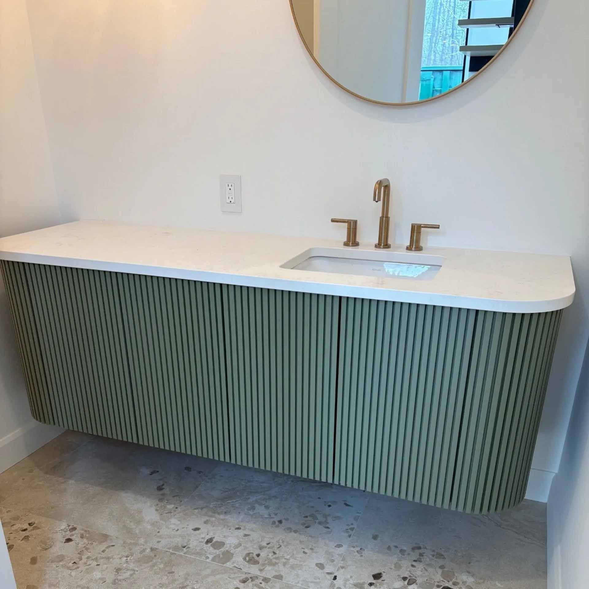 Un meuble-lavabo mural avec des portes à lattes vertes, un plan de travail blanc, un robinet en laiton et un miroir rond à cadre doré.