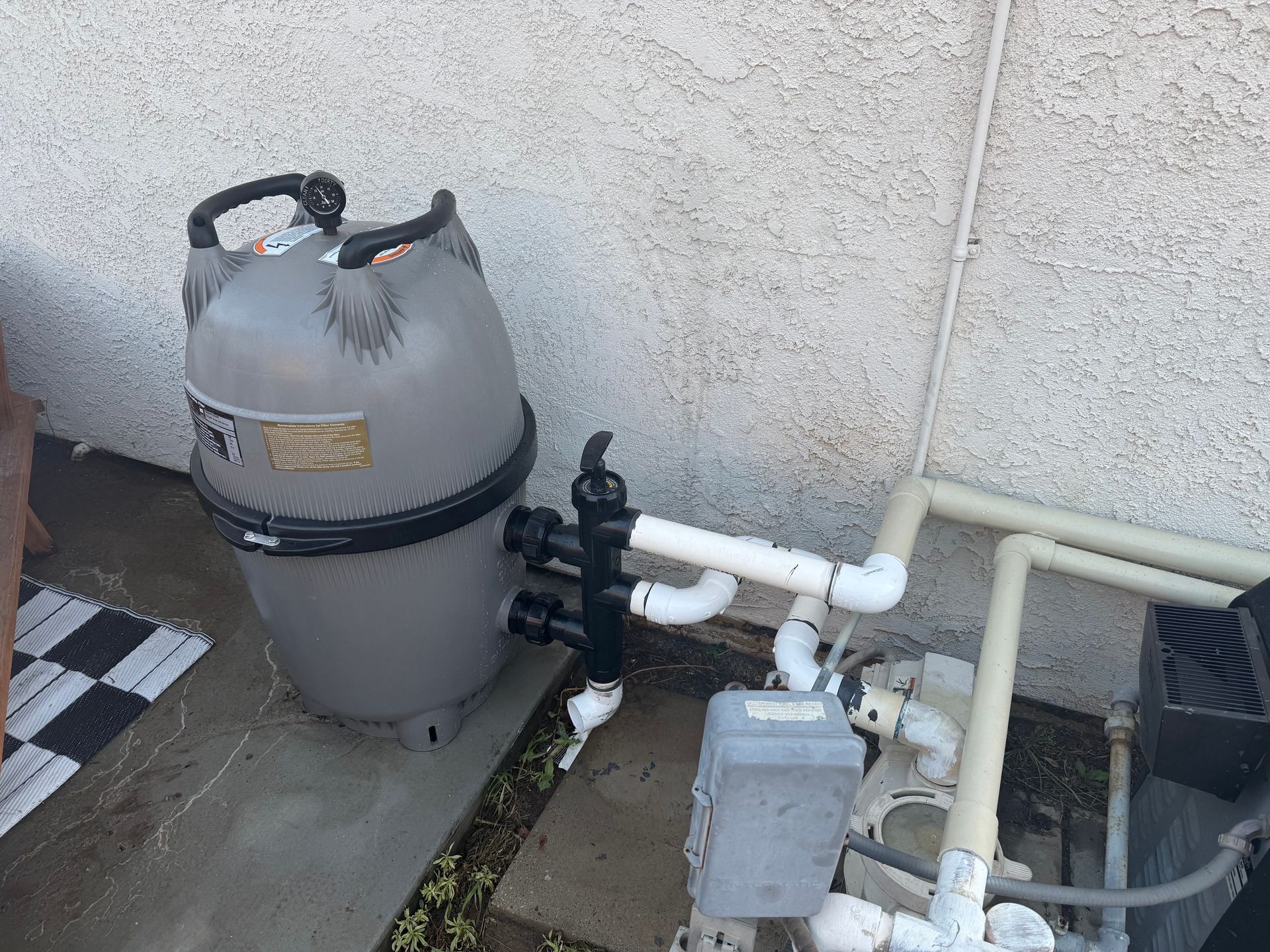 Pool filtration equipment beside a wall, with gray tank, white pipes, and control box outdoors