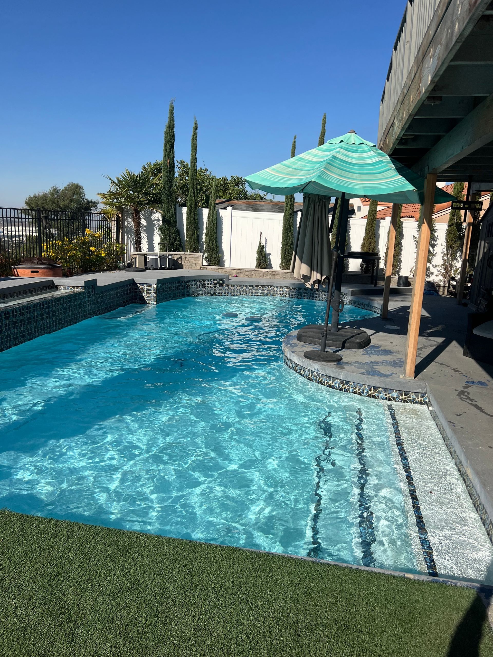 A turquoise-water pool with steps, a green umbrella, and fake grass; a sunny day in an outdoor setting.
