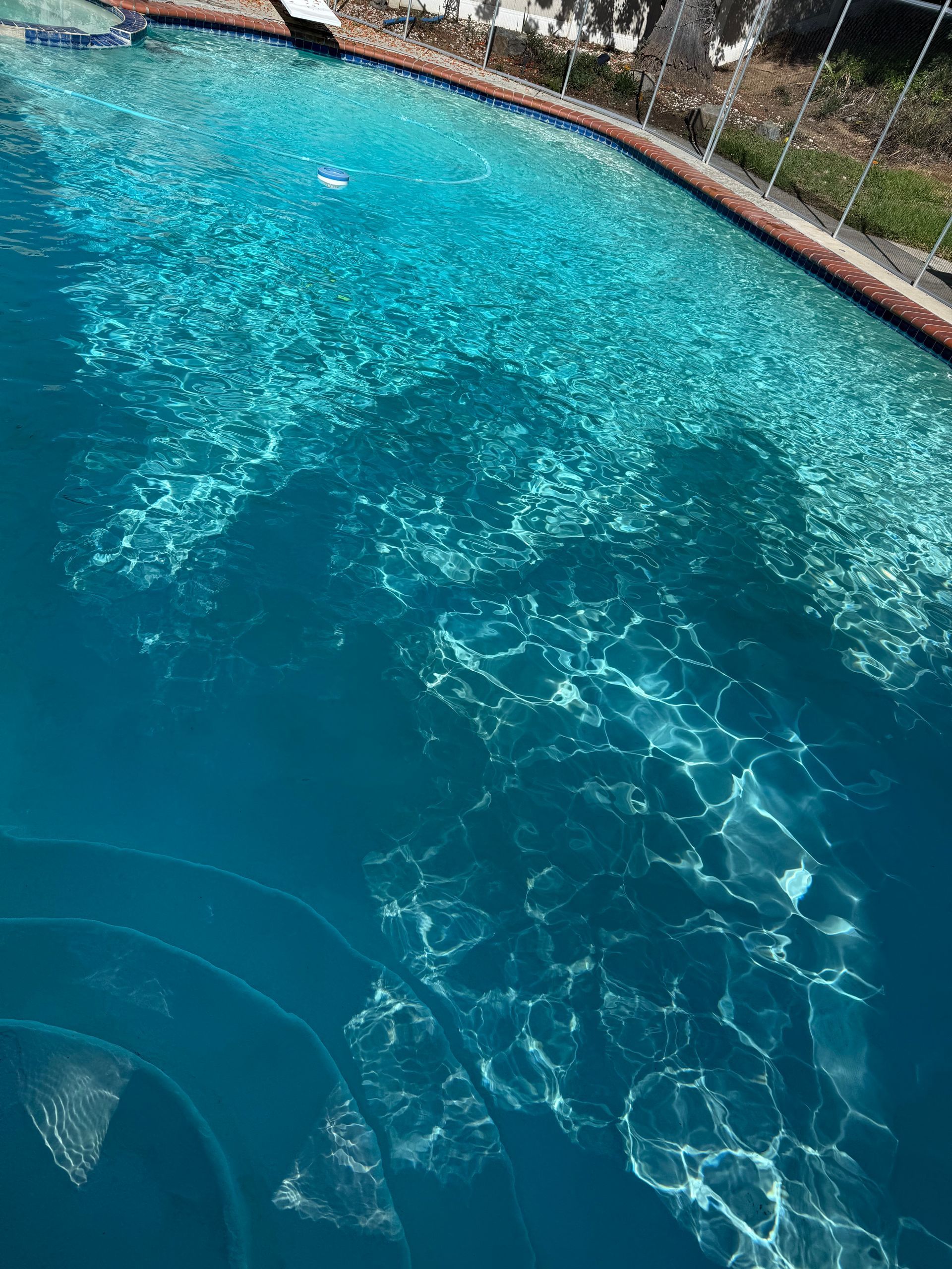 Clear blue swimming pool, sun reflecting on the water's surface, with steps leading into the pool.