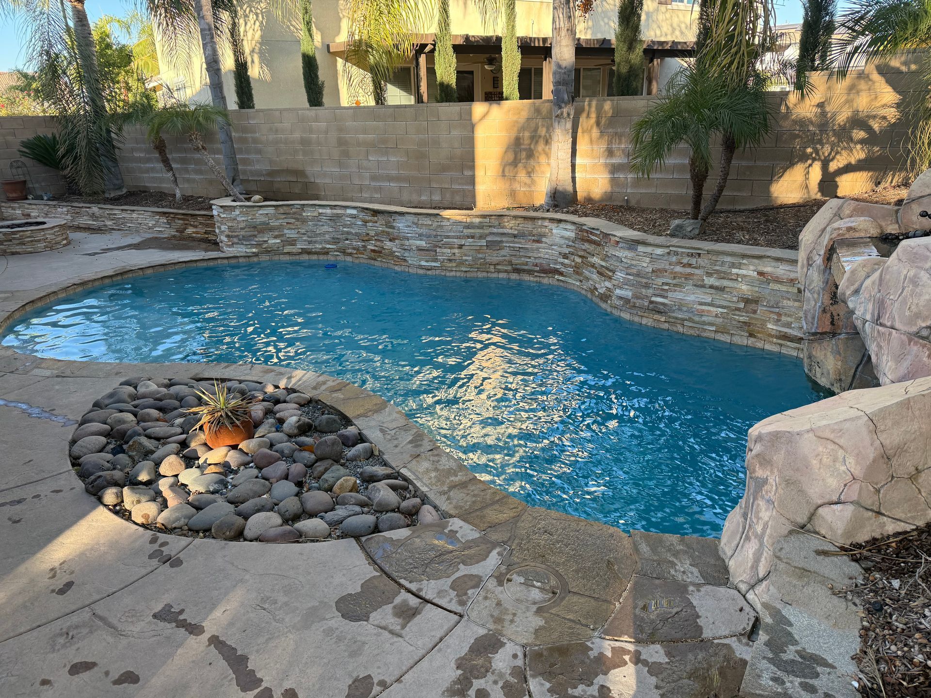 Pool with stone features, rocks, and blue water, surrounded by concrete patio and a stone wall.