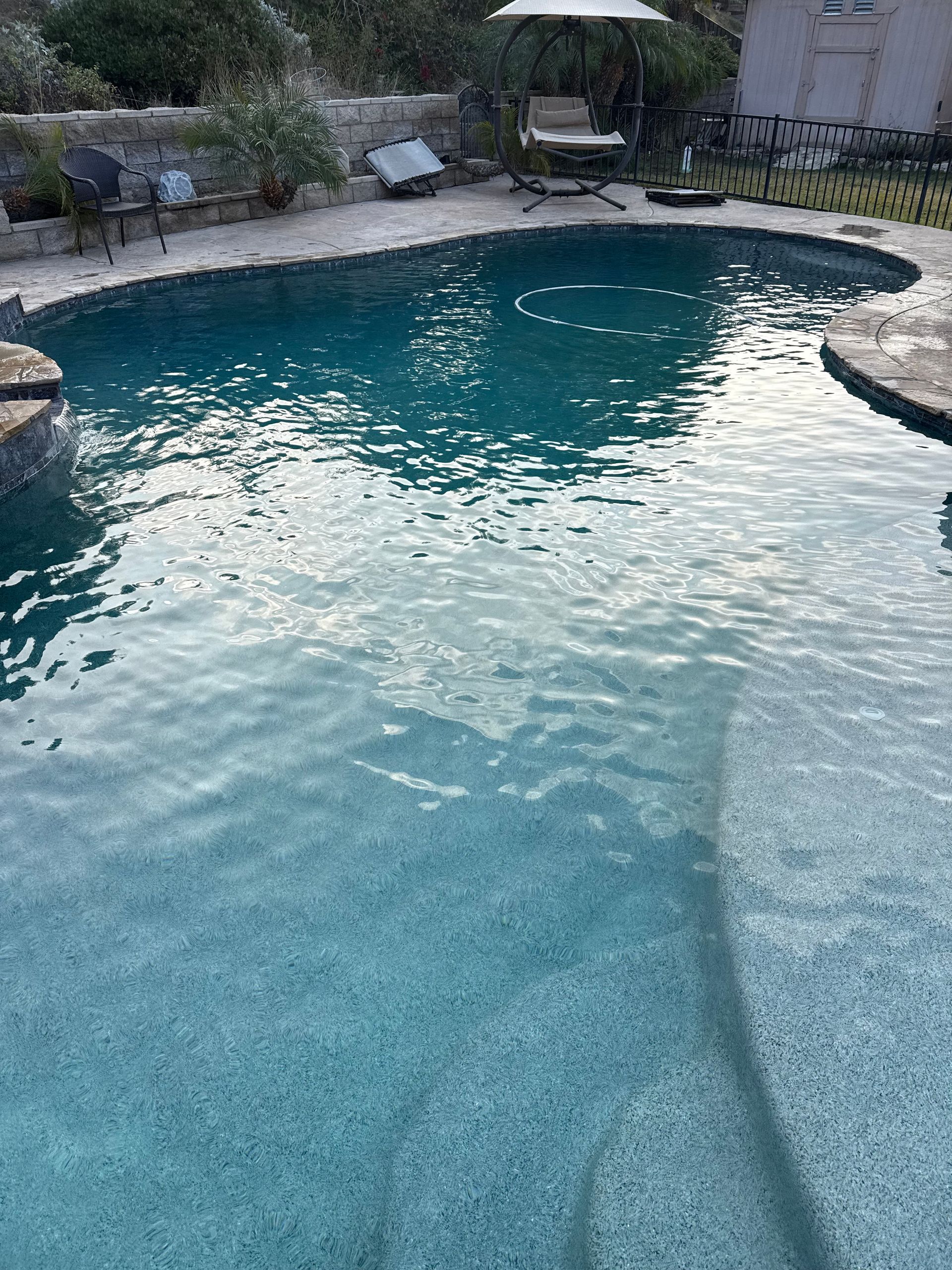 A sparkling blue swimming pool with a stone deck. Patio furniture and greenery are visible in the background.