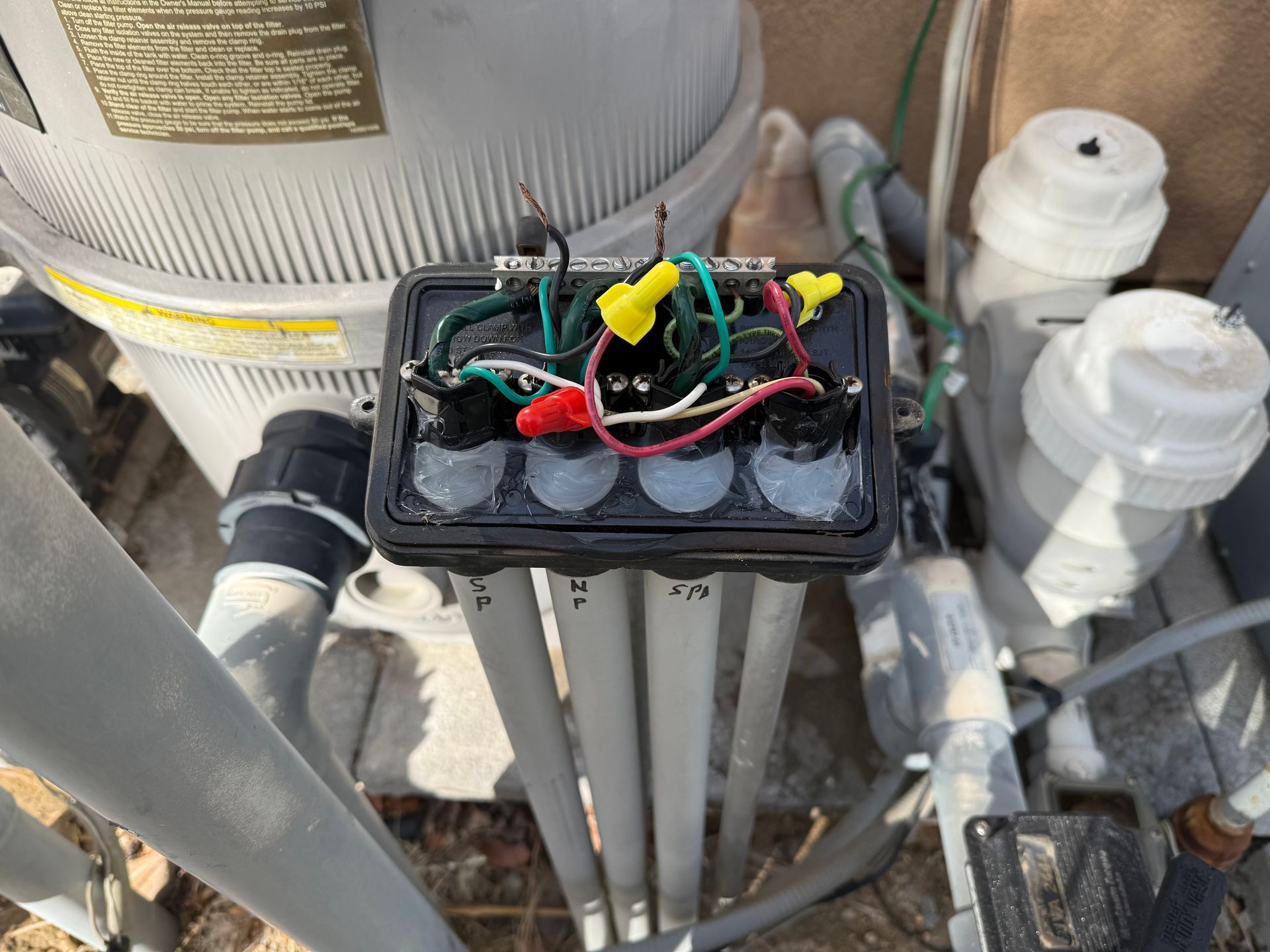 Black electrical junction box with wires and connectors, near pool equipment. Gray pipes and a white filter are also visible.