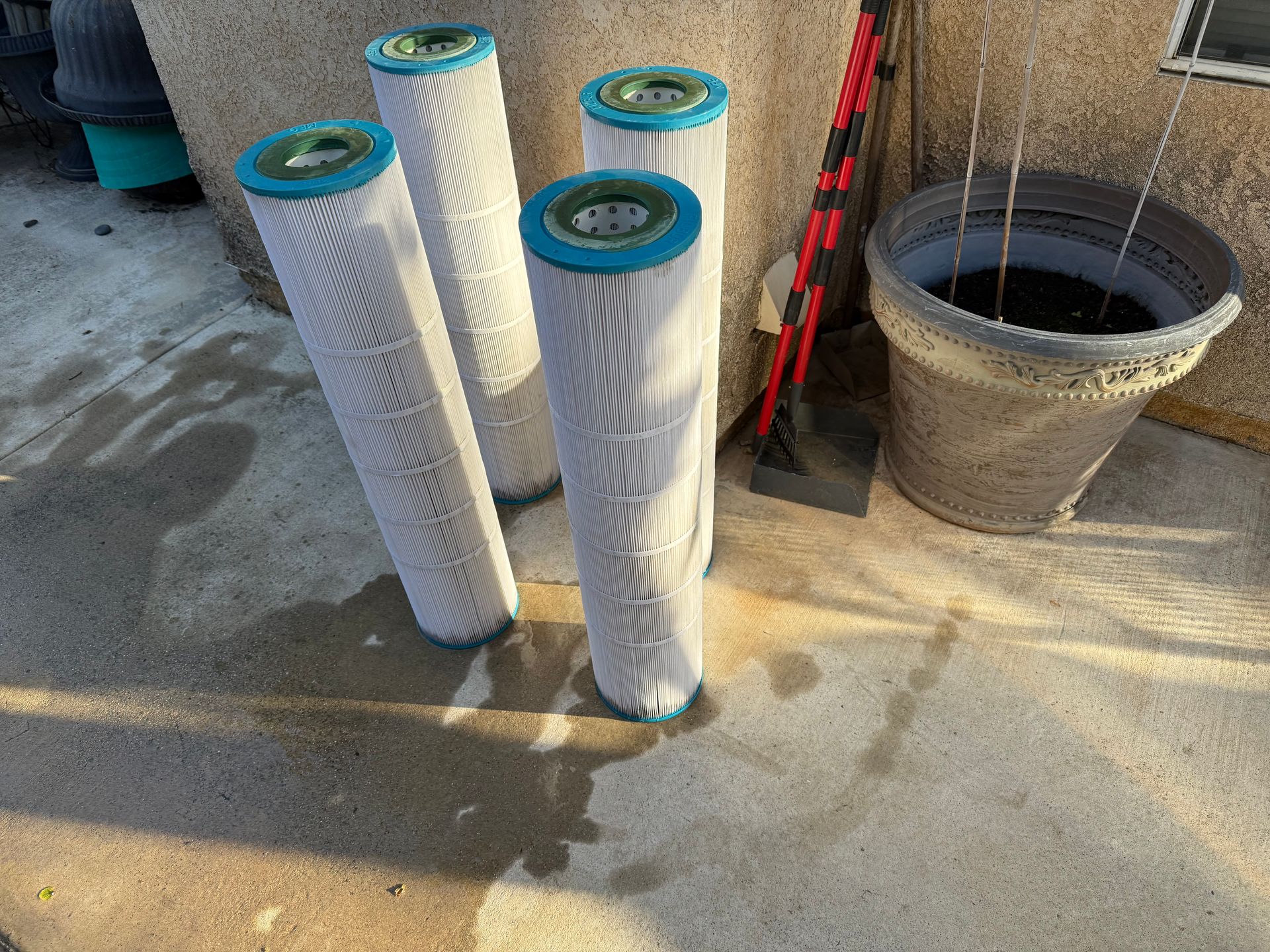Four upright pool filters with blue and green tops stand on a concrete surface next to a potted plant.