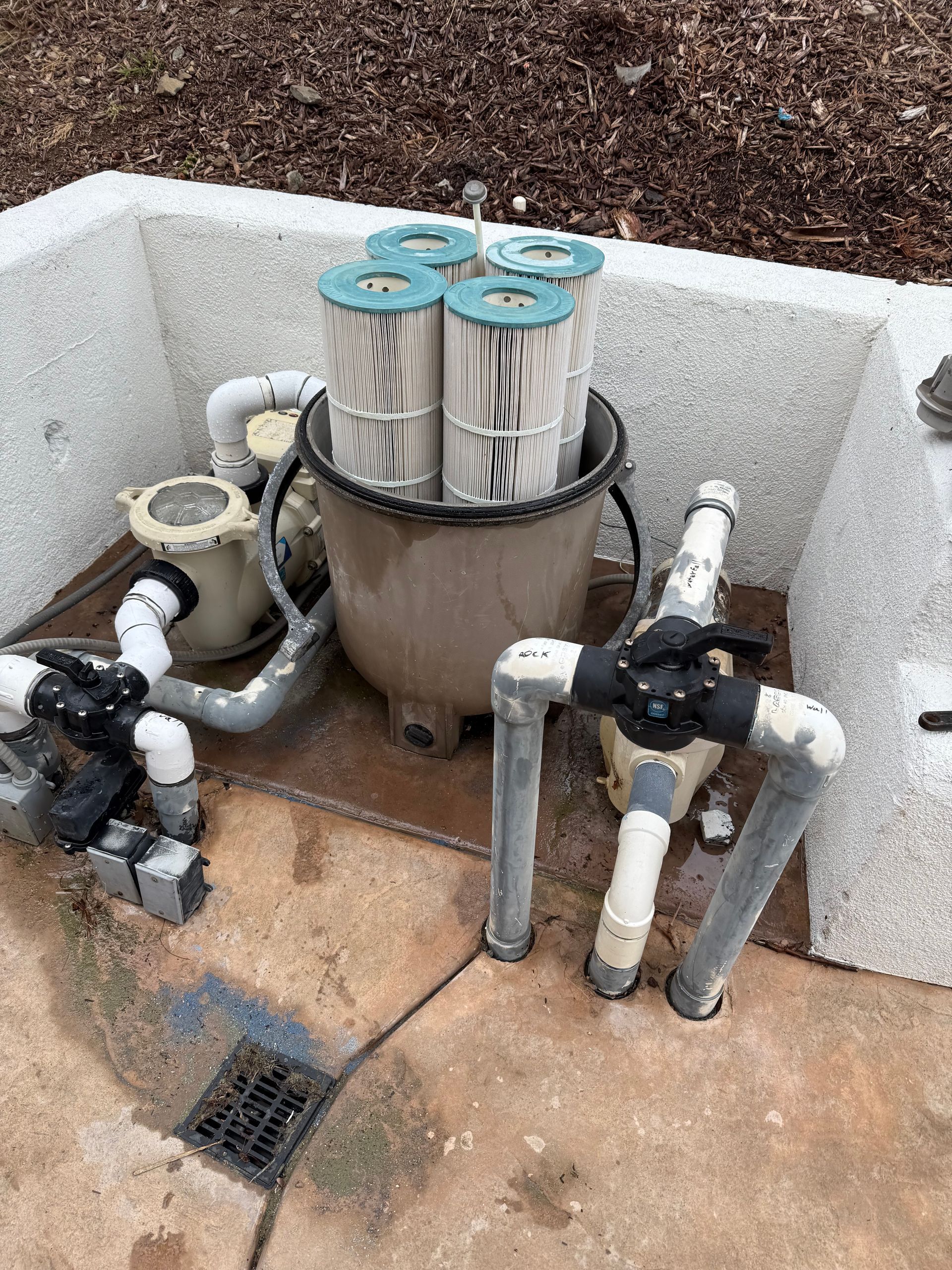 Pool filter system with multiple cartridge filters, plumbing, and valves in a concrete enclosure. The filters are beige and white.