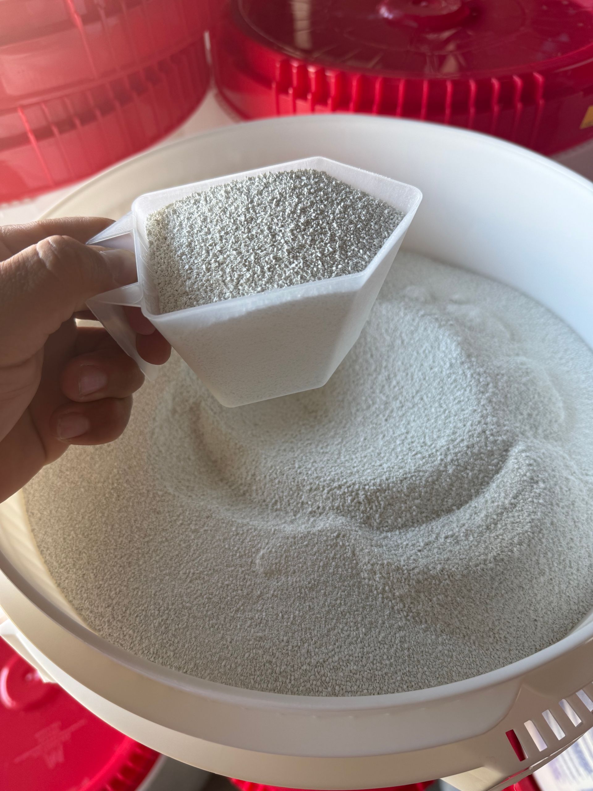 Hand holding a scoop of granular material over a white bowl filled with the same material.