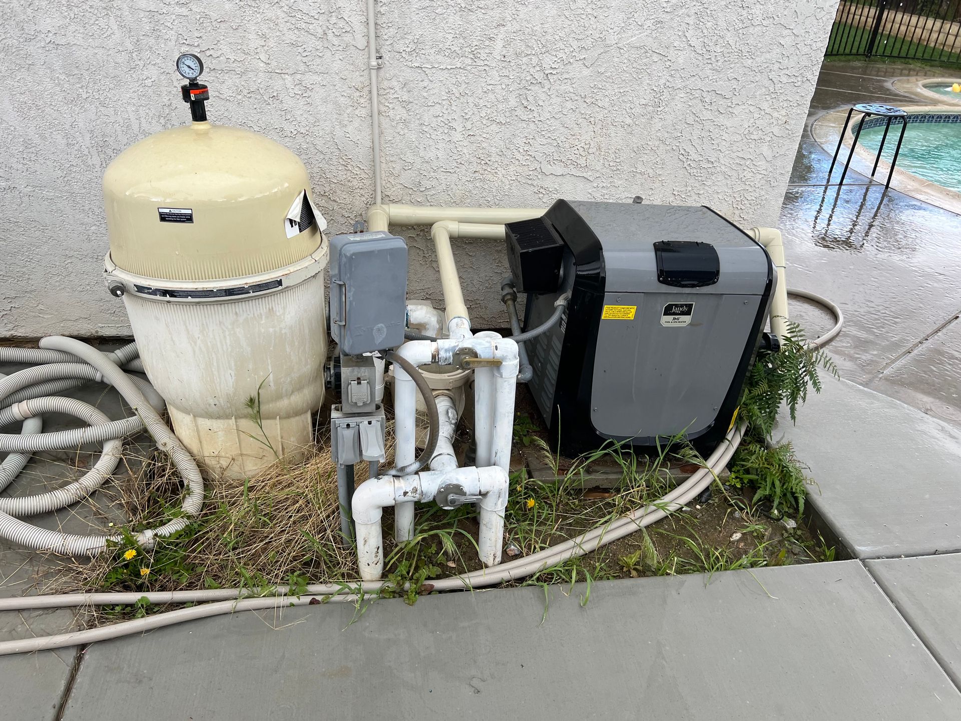 Pool equipment: a old filter, pump, and heater next to a pool. The equipment is surrounded by weeds.