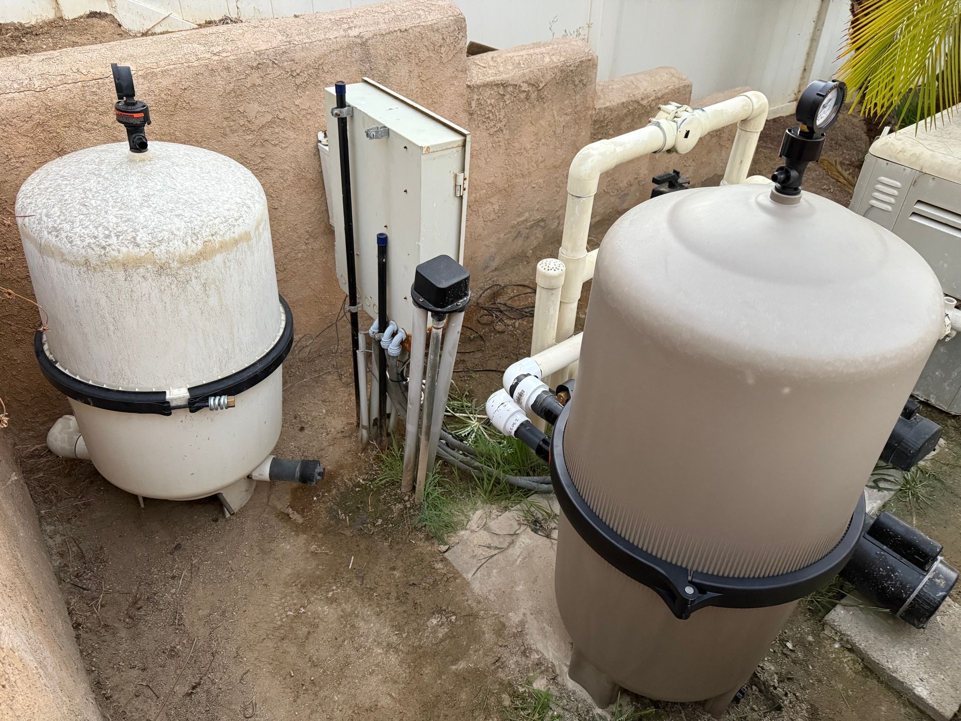 Outdoor pool filtration equipment with two large white tanks, pipes, and a control box on a concrete pad.