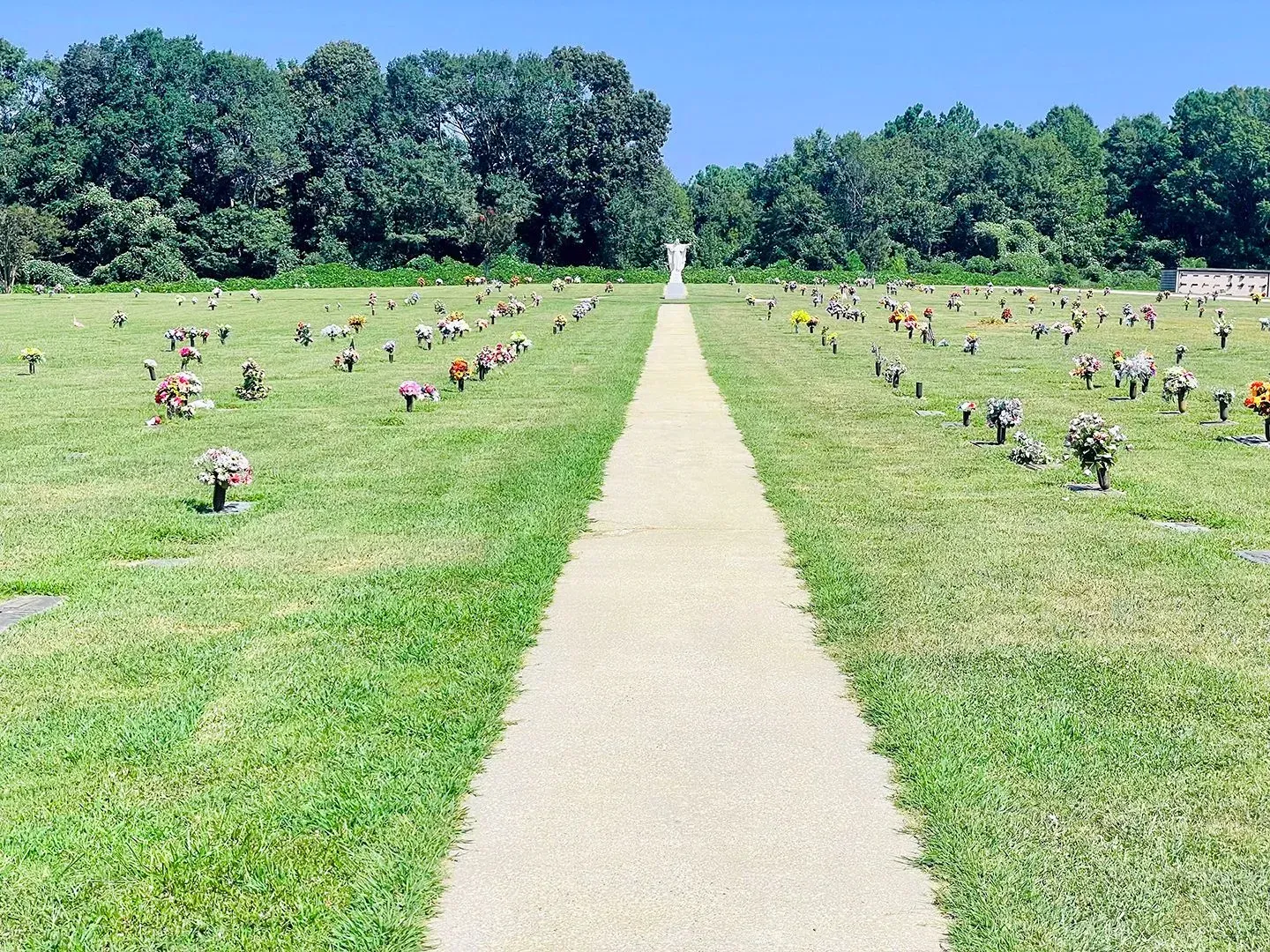 Wide cemetery with a central concrete path, rows of small headstones, and trees under a clear blue sky