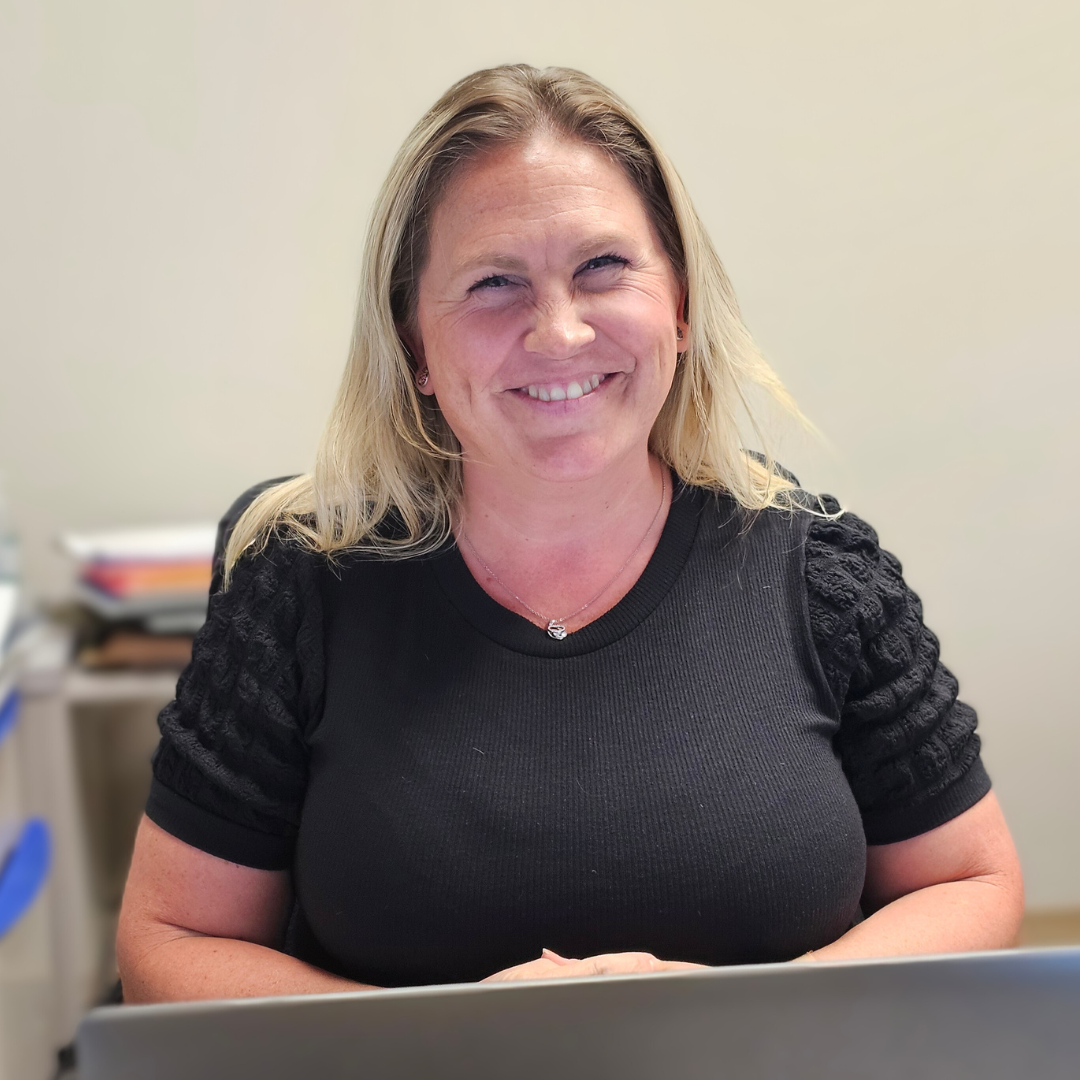 A woman is smiling while sitting at a desk in front of a laptop computer.