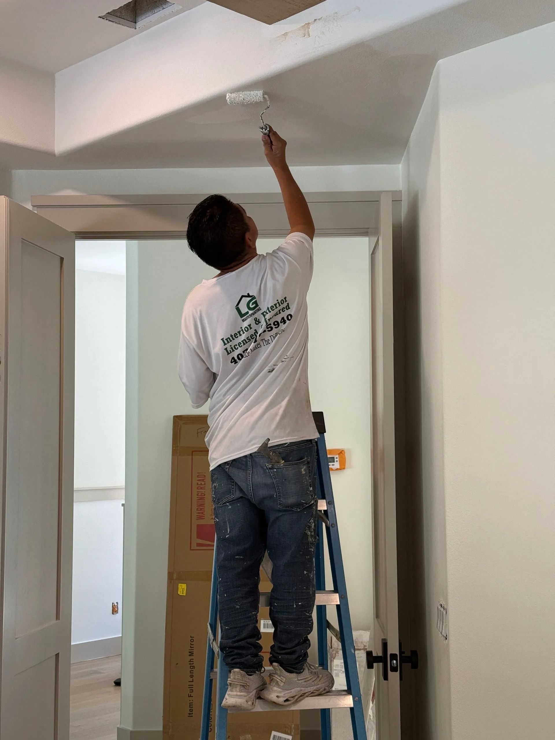 Person painting ceiling while standing on a ladder in a room with white walls and doorways.