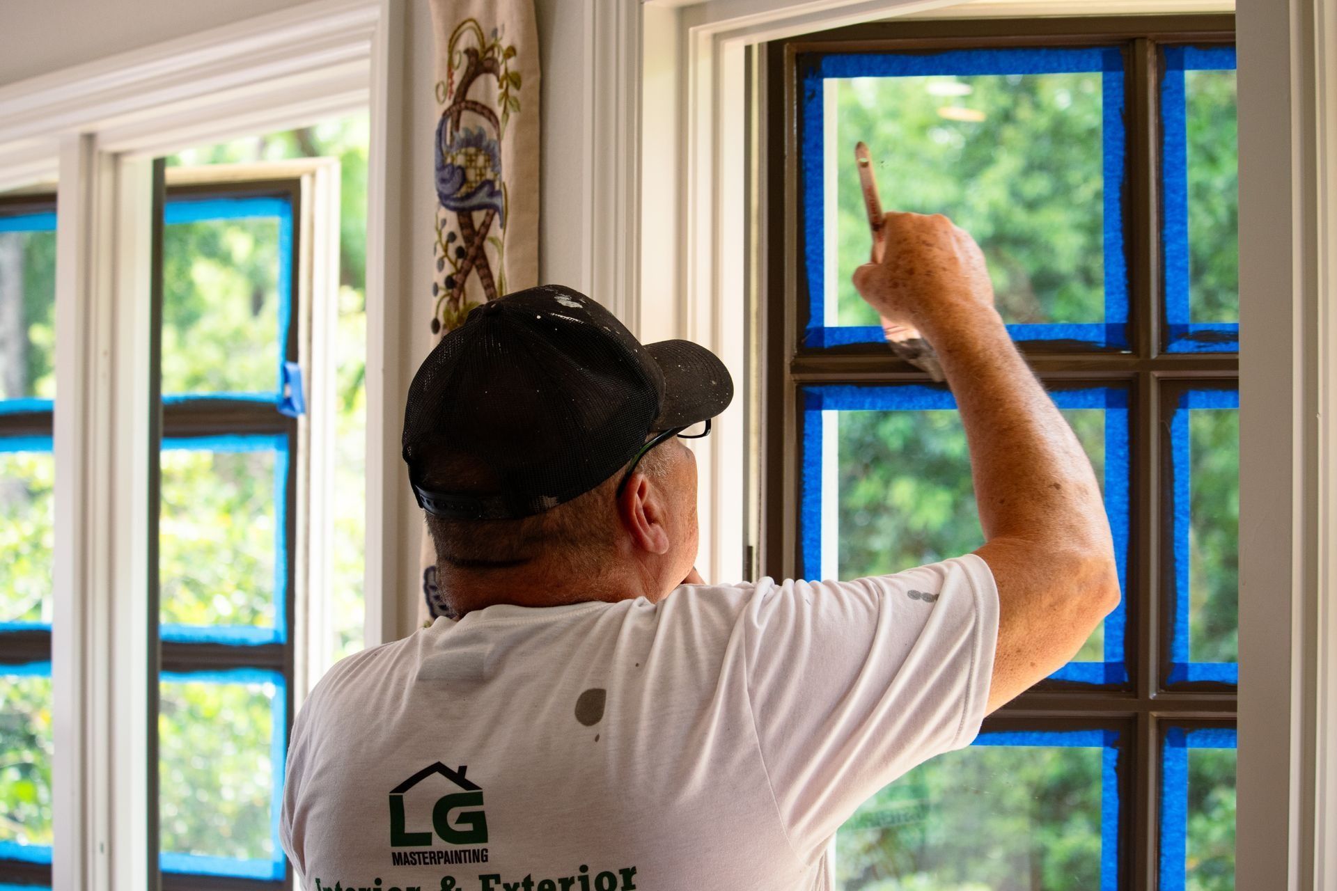 Man painting a window frame, blue tape on glass, wearing a cap and white shirt.
