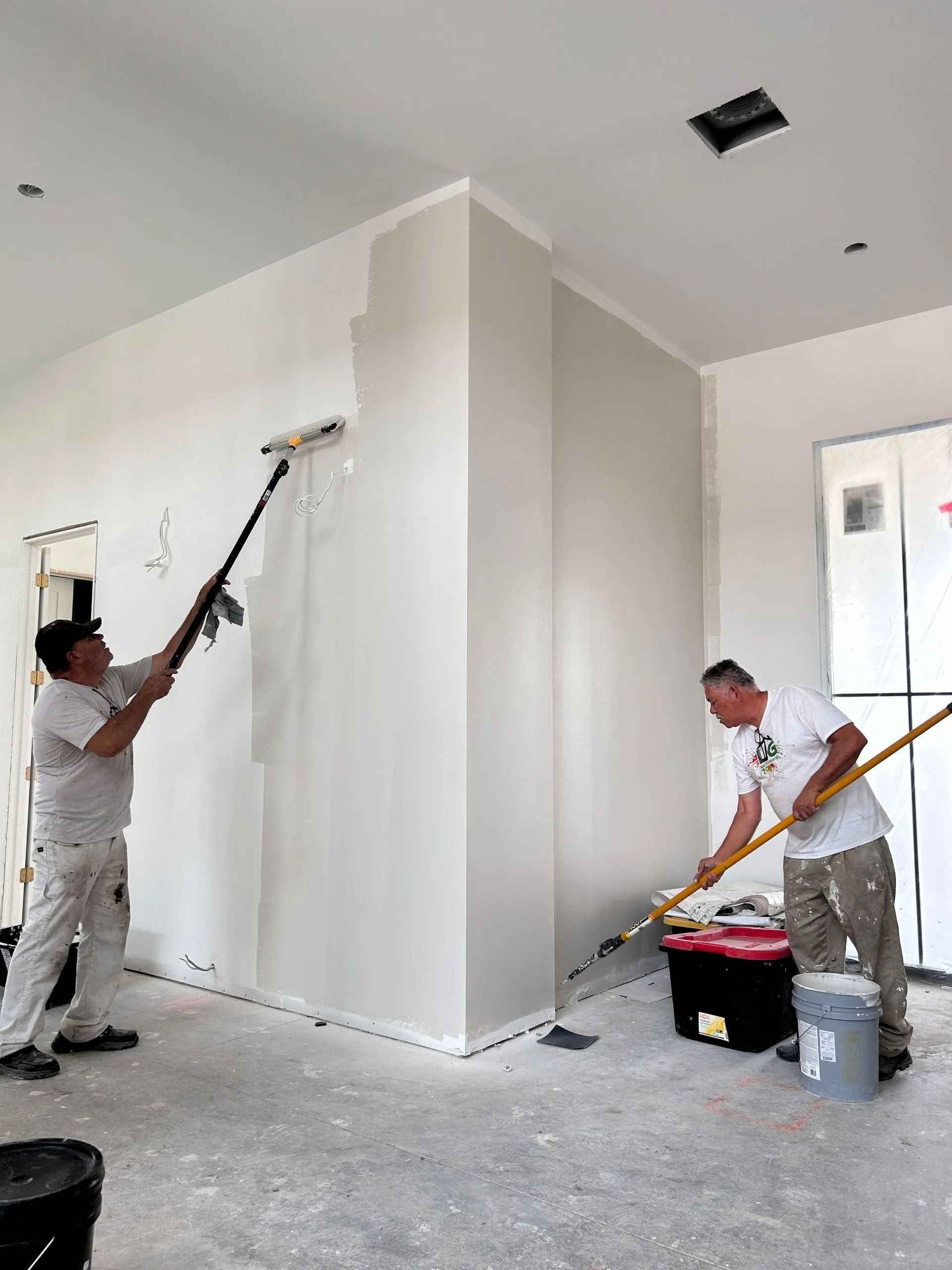 Two painters in a room painting walls with long-handled rollers. Walls are light grey, interior setting.