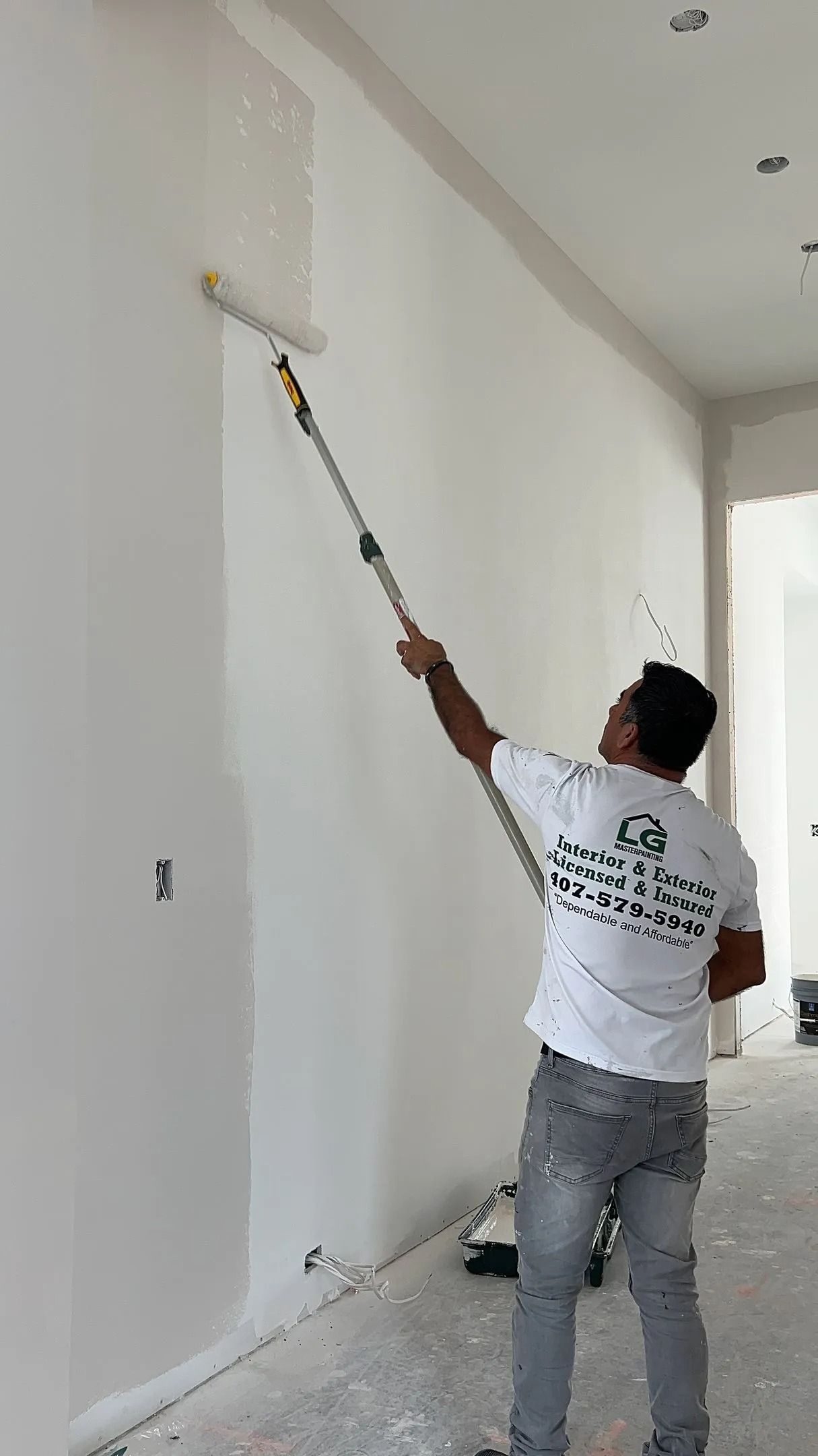 A man paints a white wall with a roller on an extension pole. He is wearing a white t-shirt and jeans.